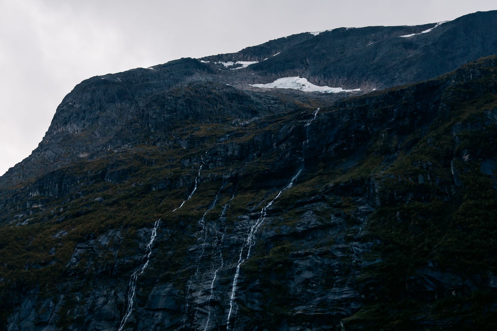 Thin waterfalls running the full height of a dark mountainside from a remnant snowfield