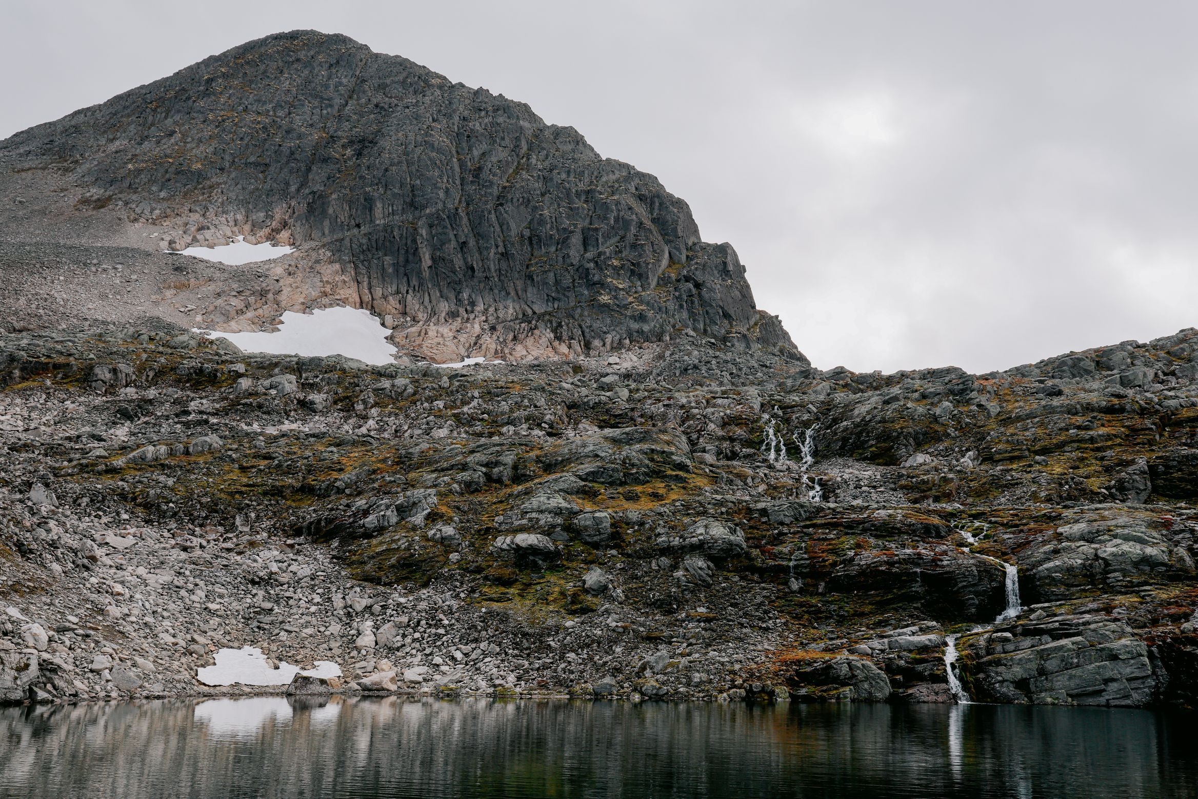 A waterfall feeding a black tarn beneath a rounded crag streaked with old snow