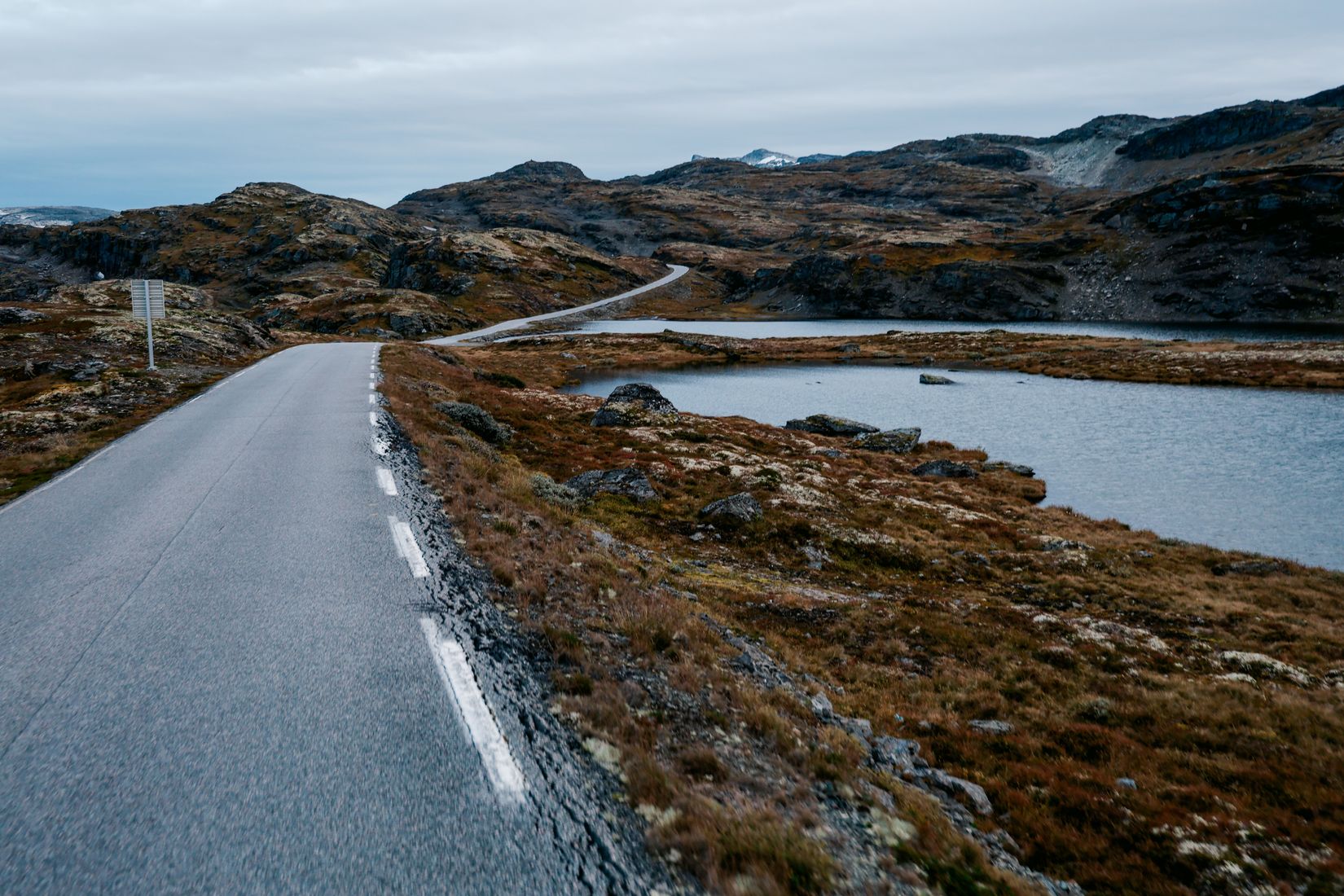 An empty road curving across a bare mountain plateau past shallow lakes
