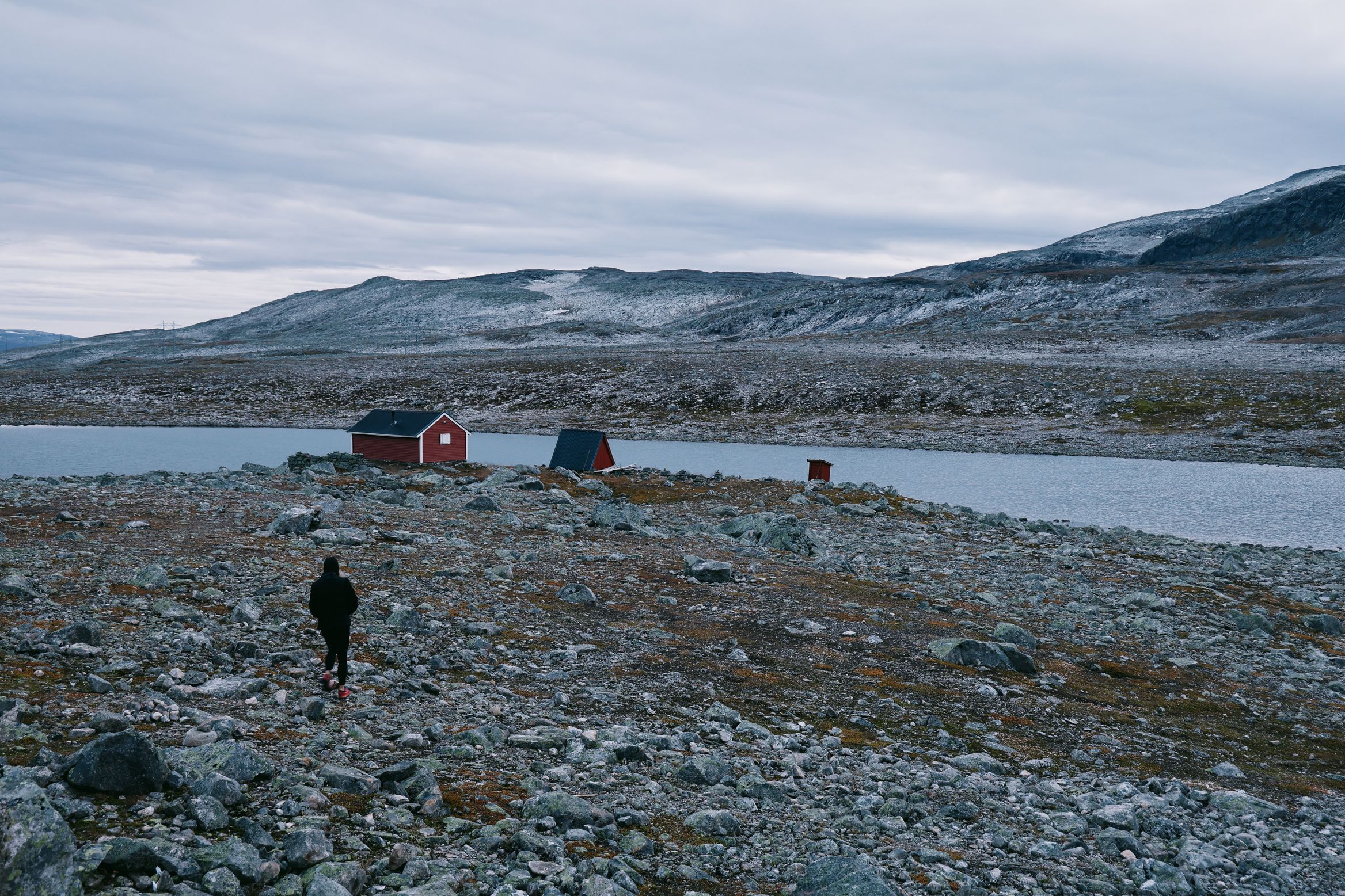 A figure walking over stony ground towards a red hut and a tent beside a high lake, snow on the hills behind