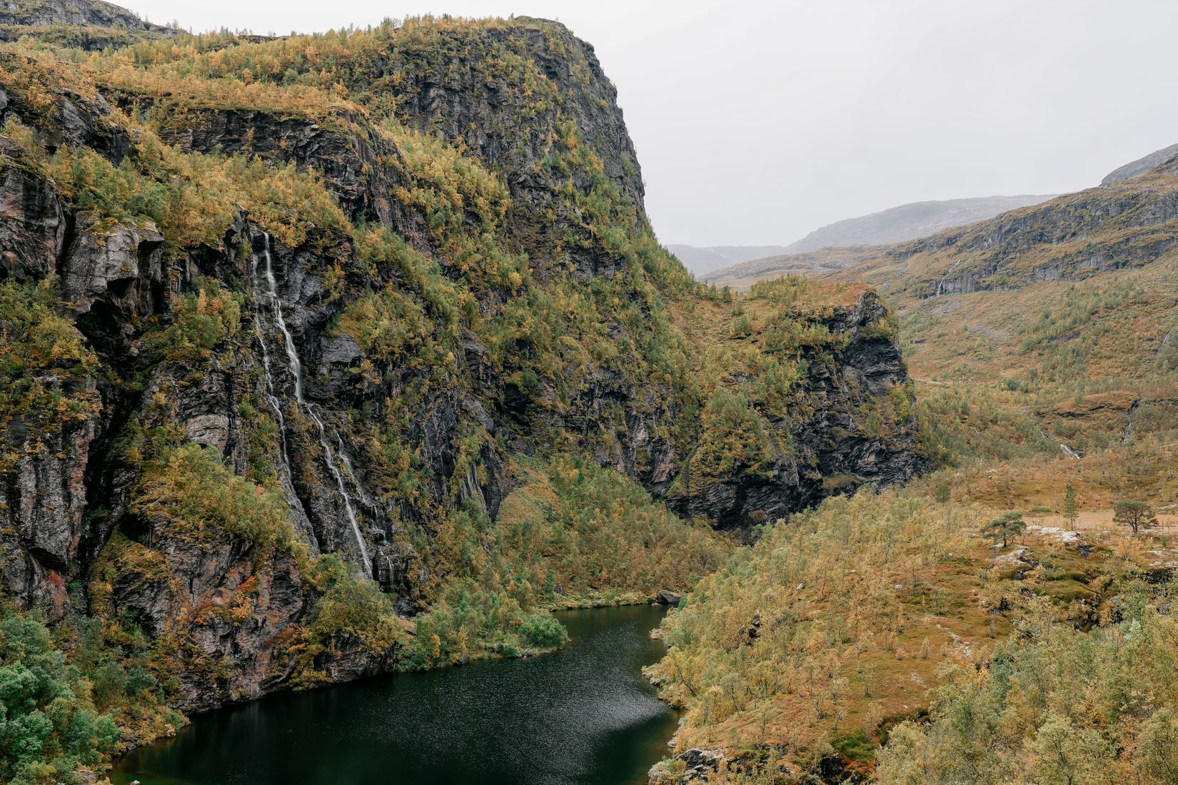 A narrow gorge lake between cliffs hung with autumn birch, a waterfall running down the left wall