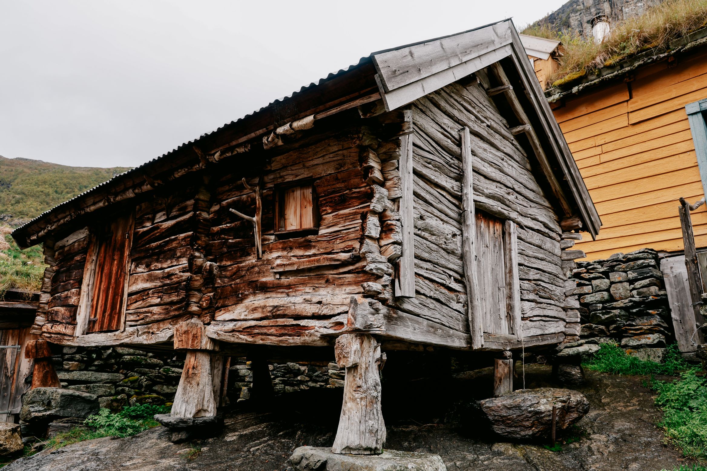 An old timber stabbur raised on stone-topped legs, a yellow farmhouse behind it