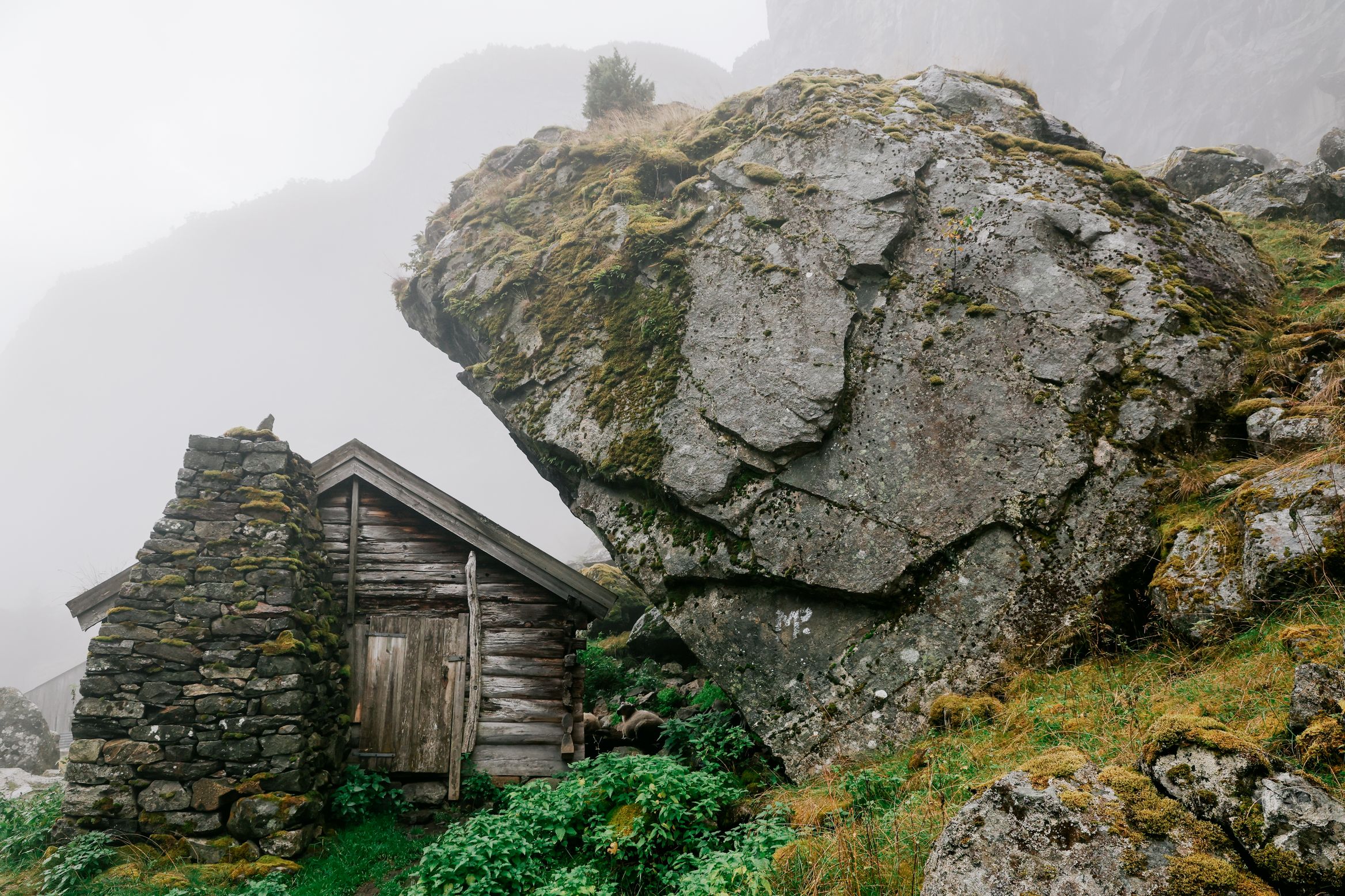A tiny stone and timber hut tucked under an enormous split boulder in the mist