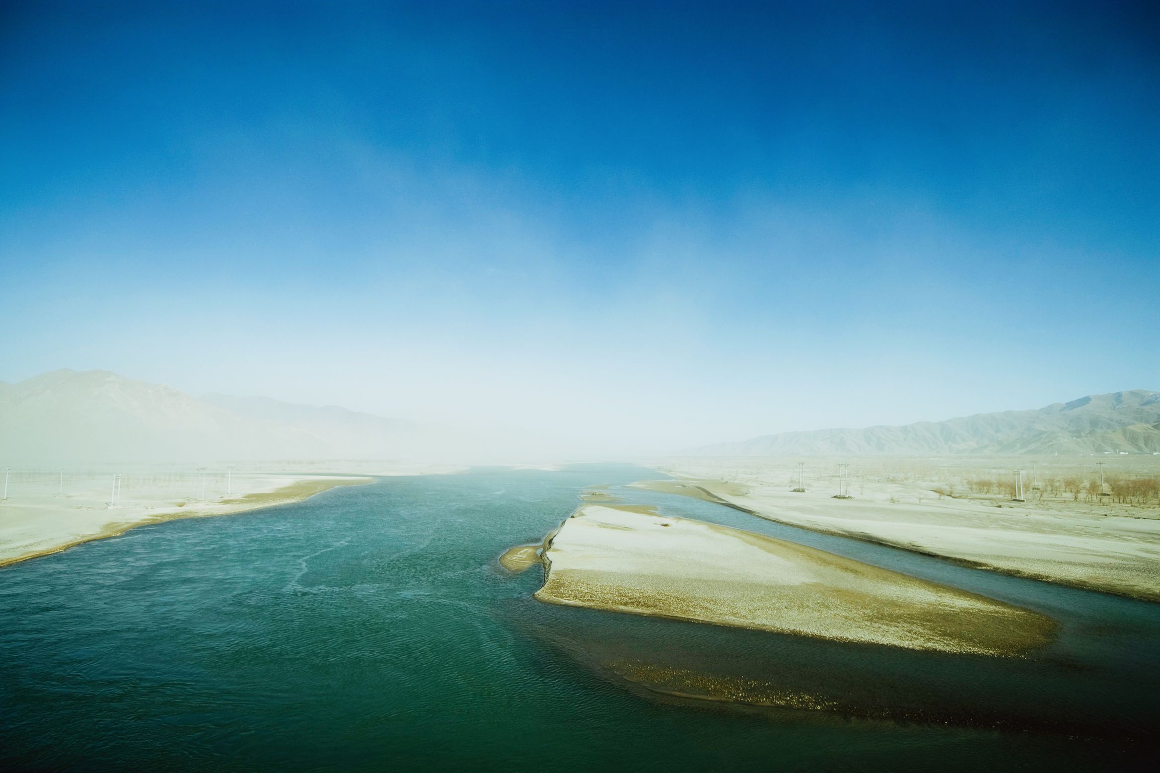 A wide braided river running between pale sandbanks, dust haze over the hills