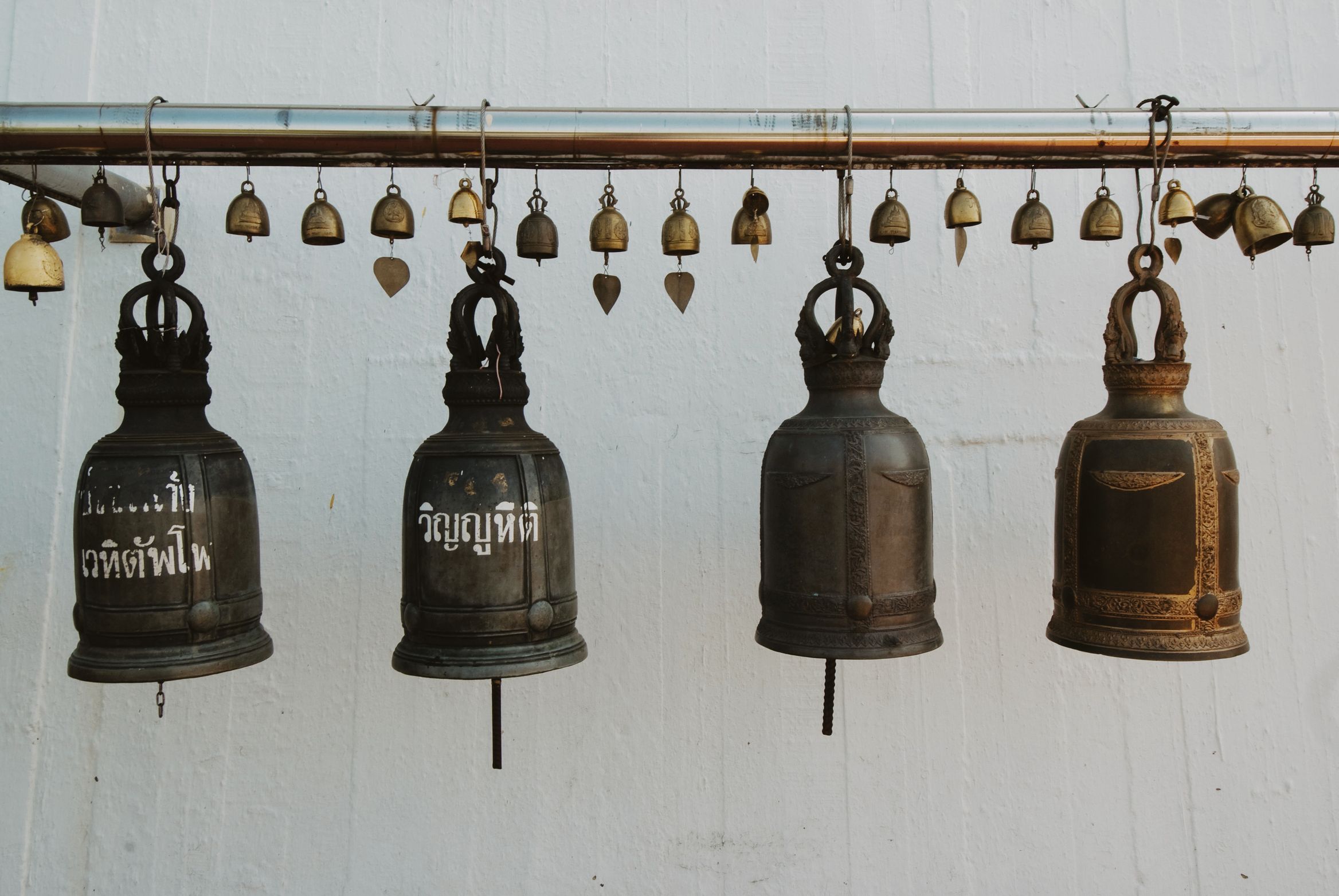Four bronze bells hung on a rail against a whitewashed wall, small bells strung above them