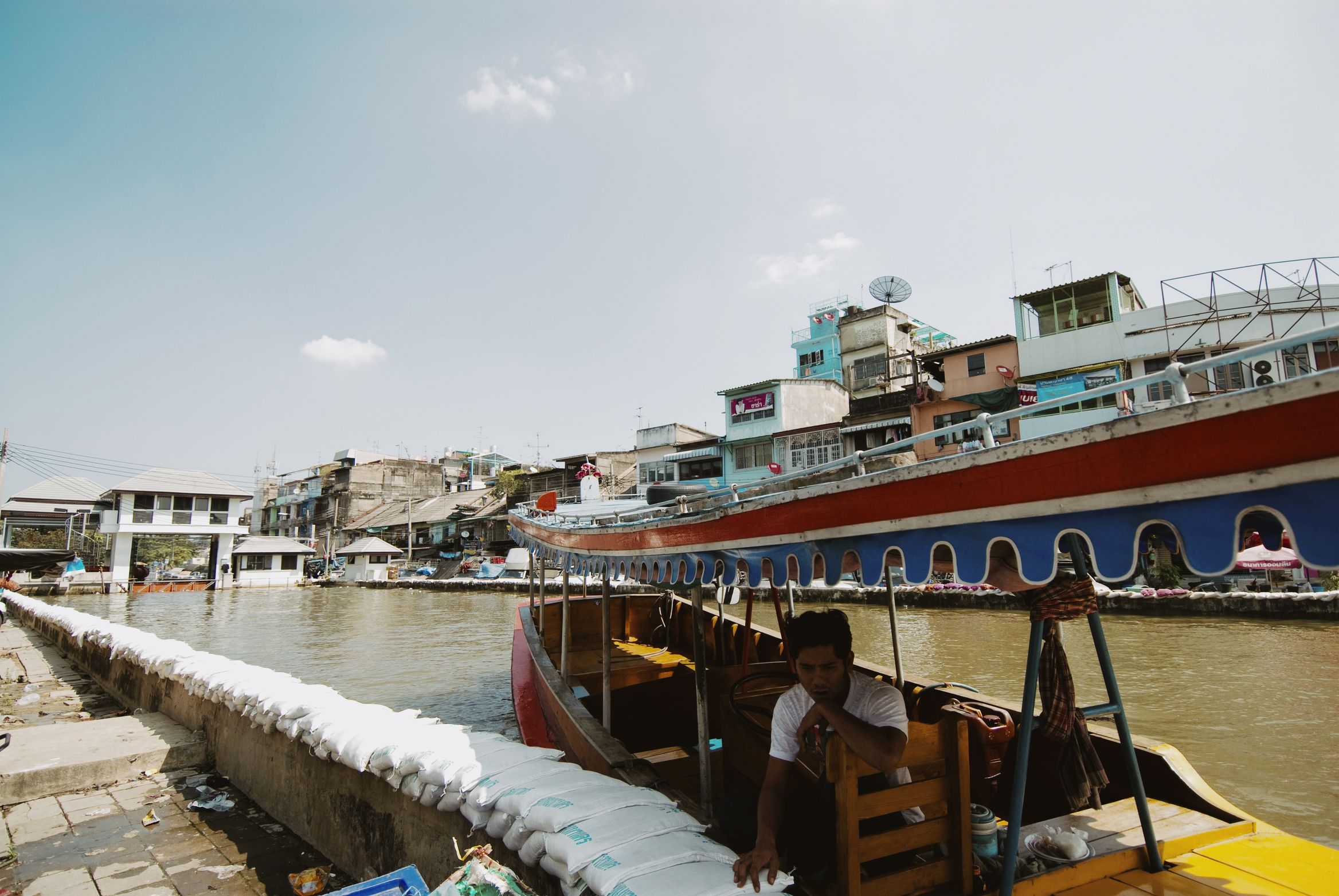 A boatman sitting at the stern of a long boat moored against a sandbagged canal wall