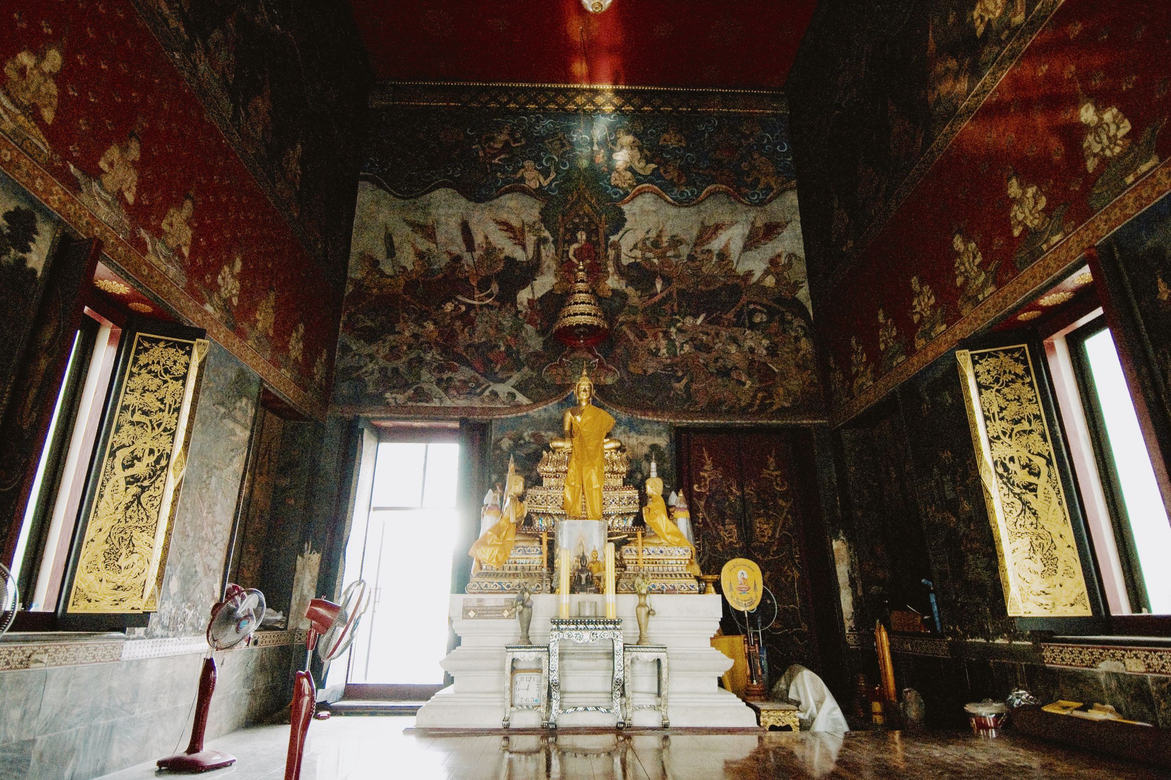 A temple hall lit from a doorway behind the standing gold Buddha, murals over every wall