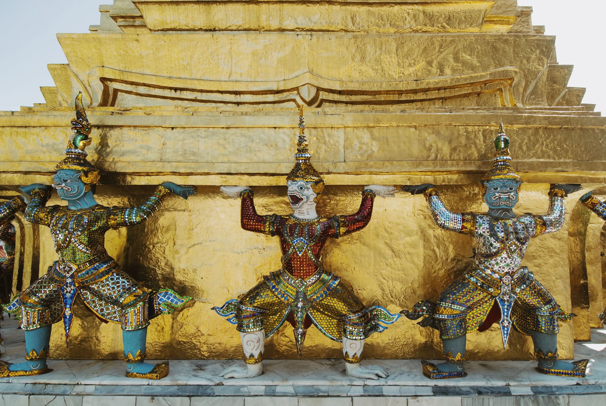 A row of mosaic demon figures holding up the gold base of a chedi