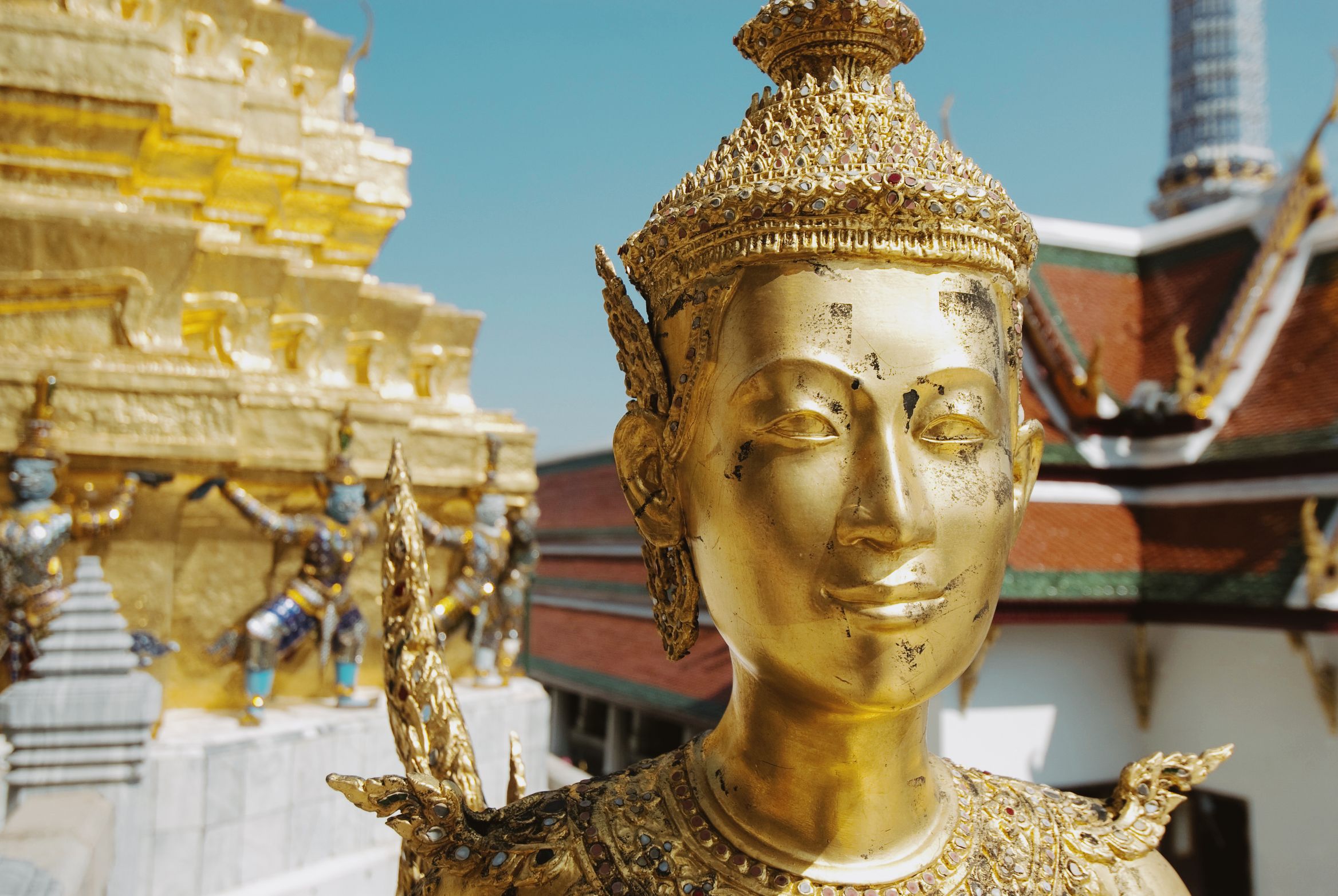 The face of a gold kinnara statue in full sun, temple roofs behind