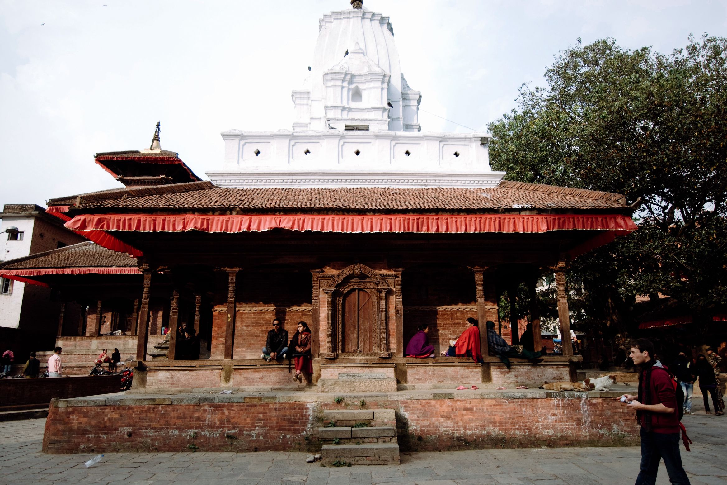 A low temple with a red cloth valance under its eaves, a white dome behind it, people resting on the platform