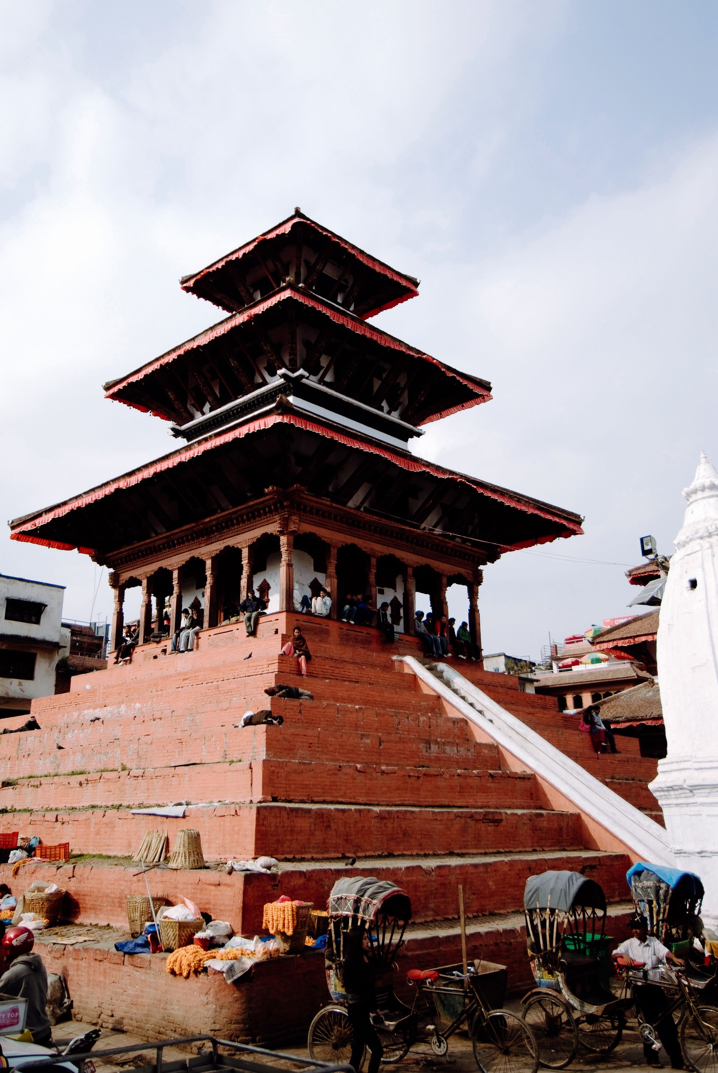 A tiered red temple rising off its stepped plinth above the square, people sitting along the top step