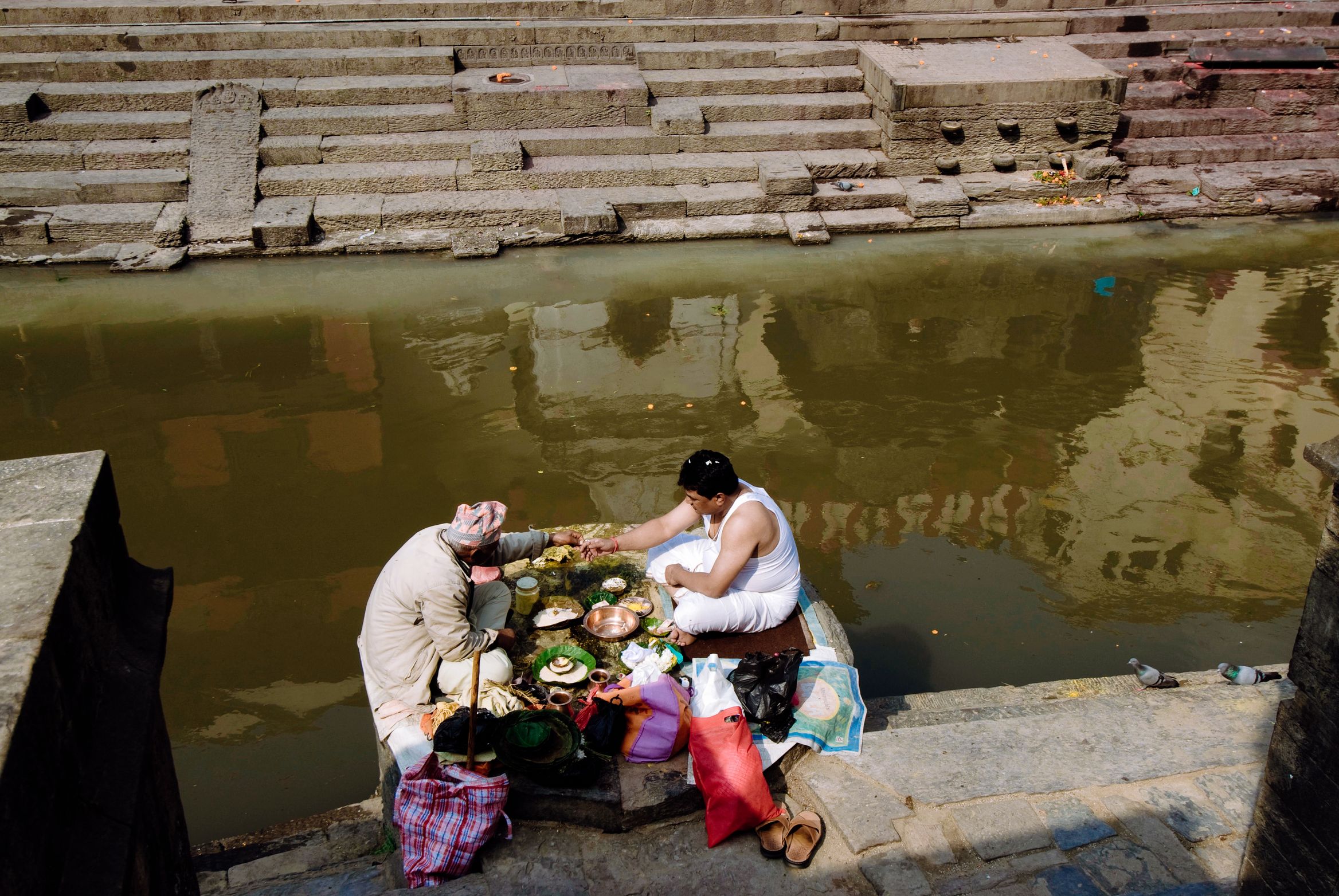 Two people laying out an offering on the ghat steps beside green river water
