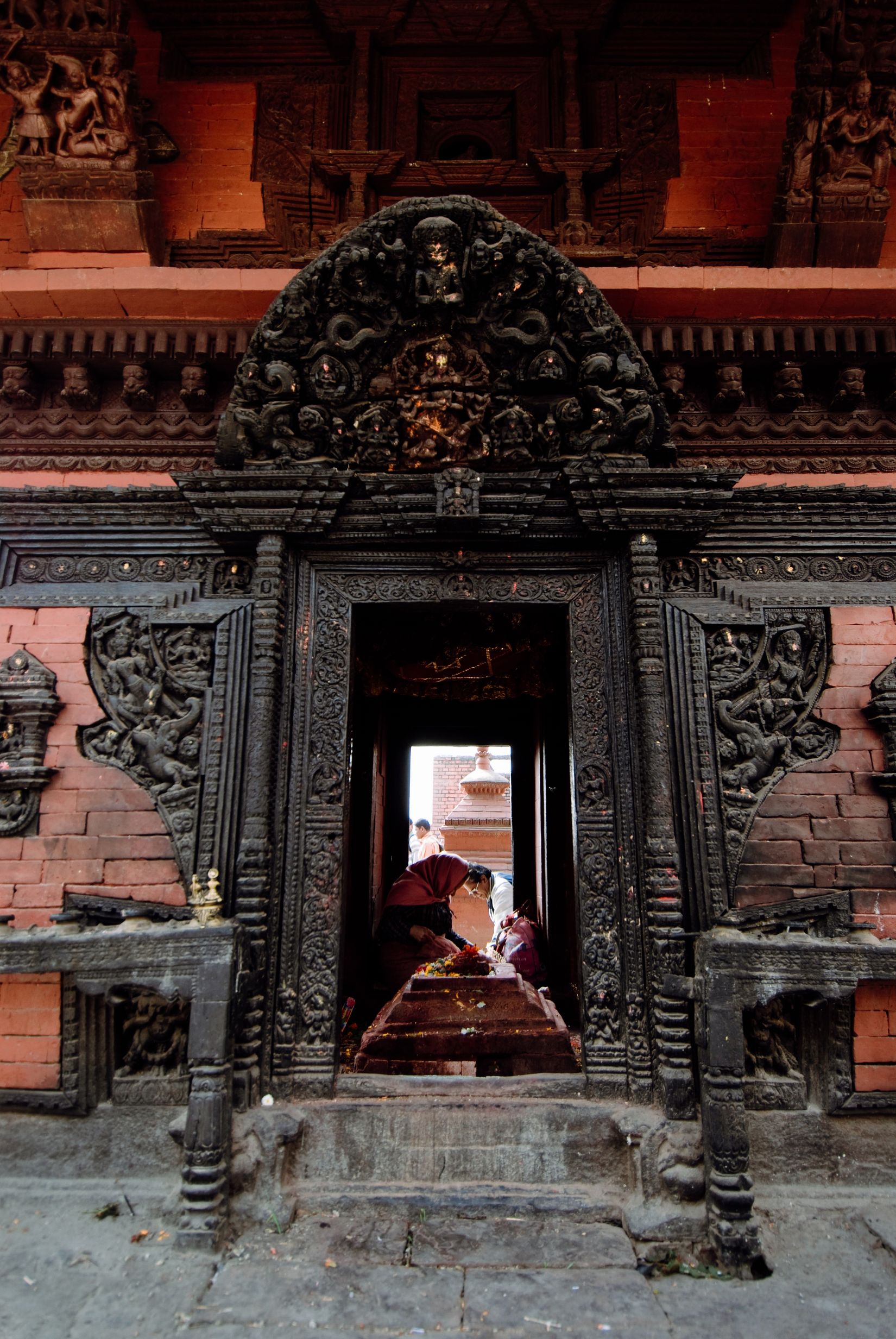 A dark carved wooden doorway in a brick wall, someone in red laying an offering on the step in the lit courtyard beyond