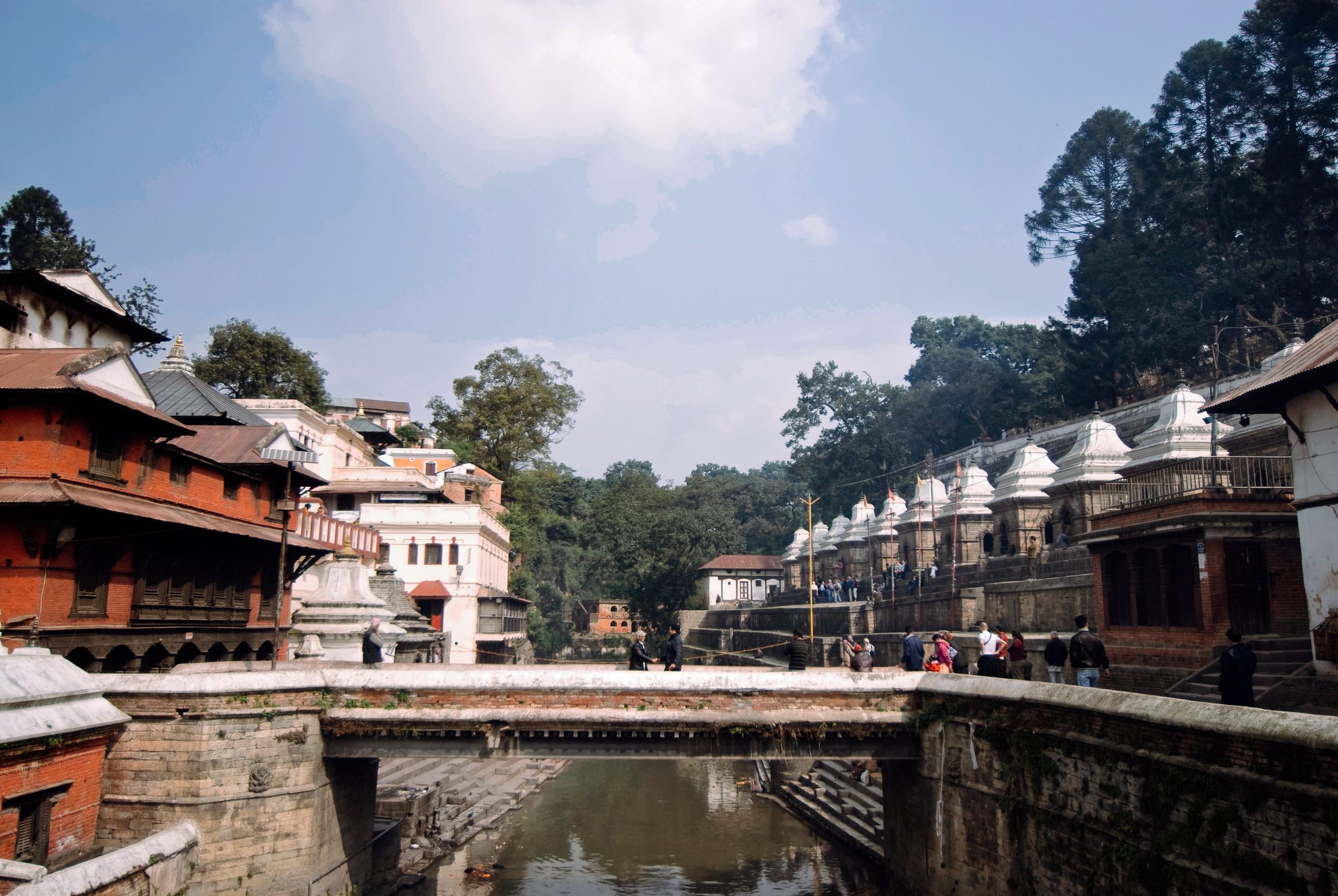 A bridge over the low river, temple roofs on one bank and a row of white shrines on the other