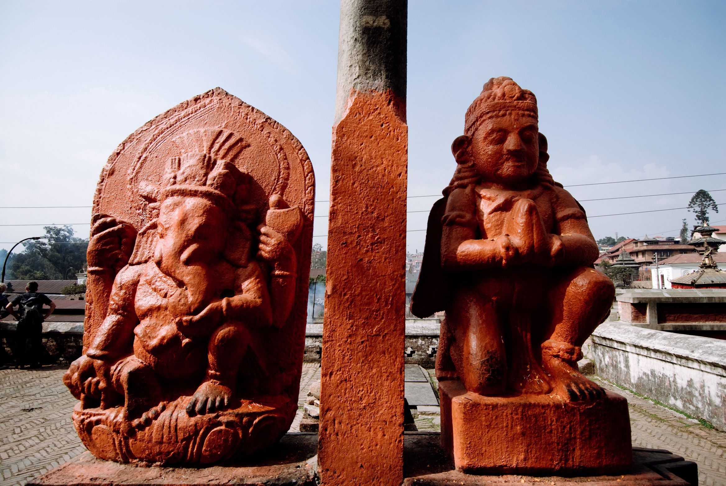 Two orange-painted stone figures either side of a post, Ganesh on the left and a kneeling Hanuman on the right
