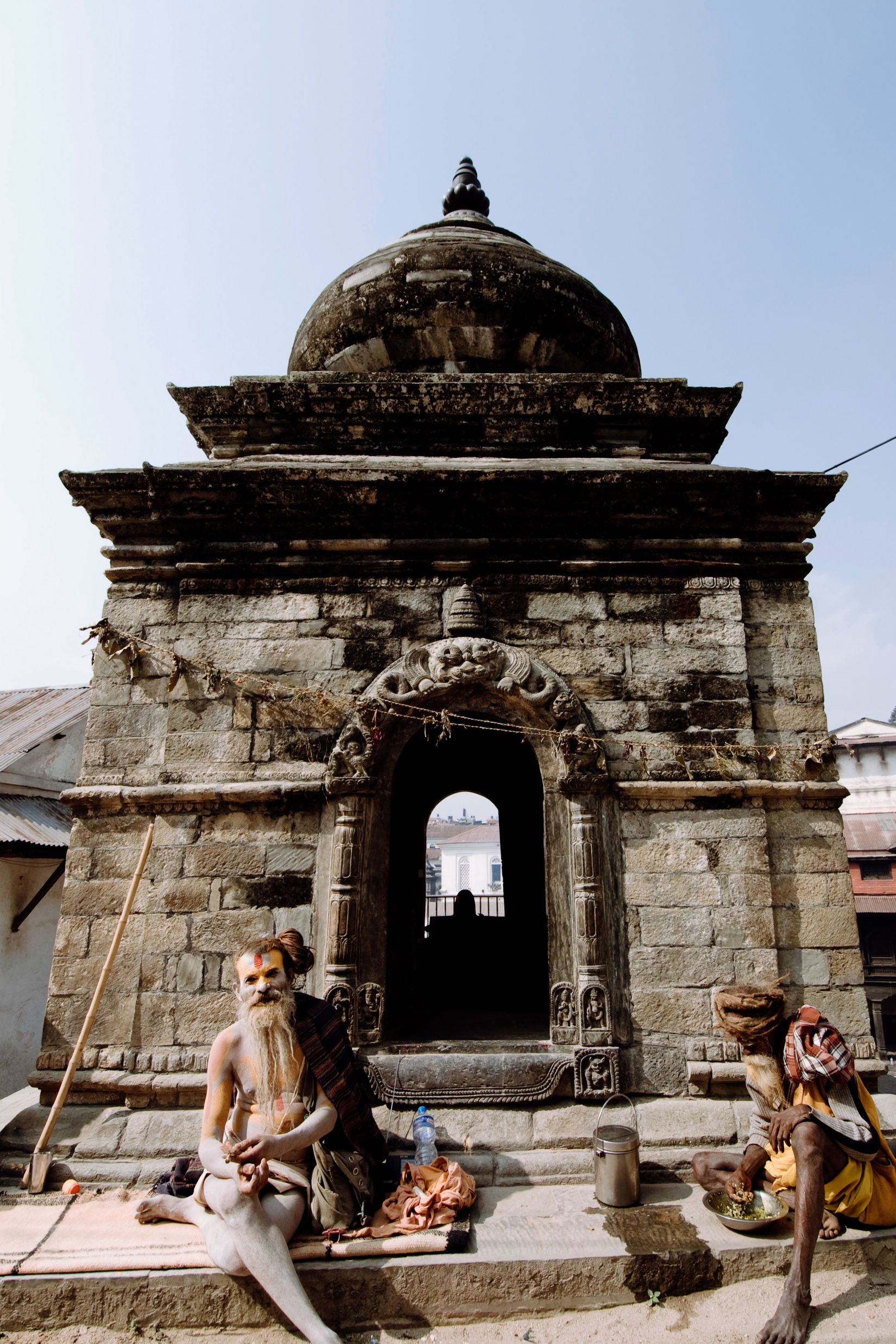 Two sadhus sitting on the steps of a stone shrine in hard midday sun, one with a long white beard