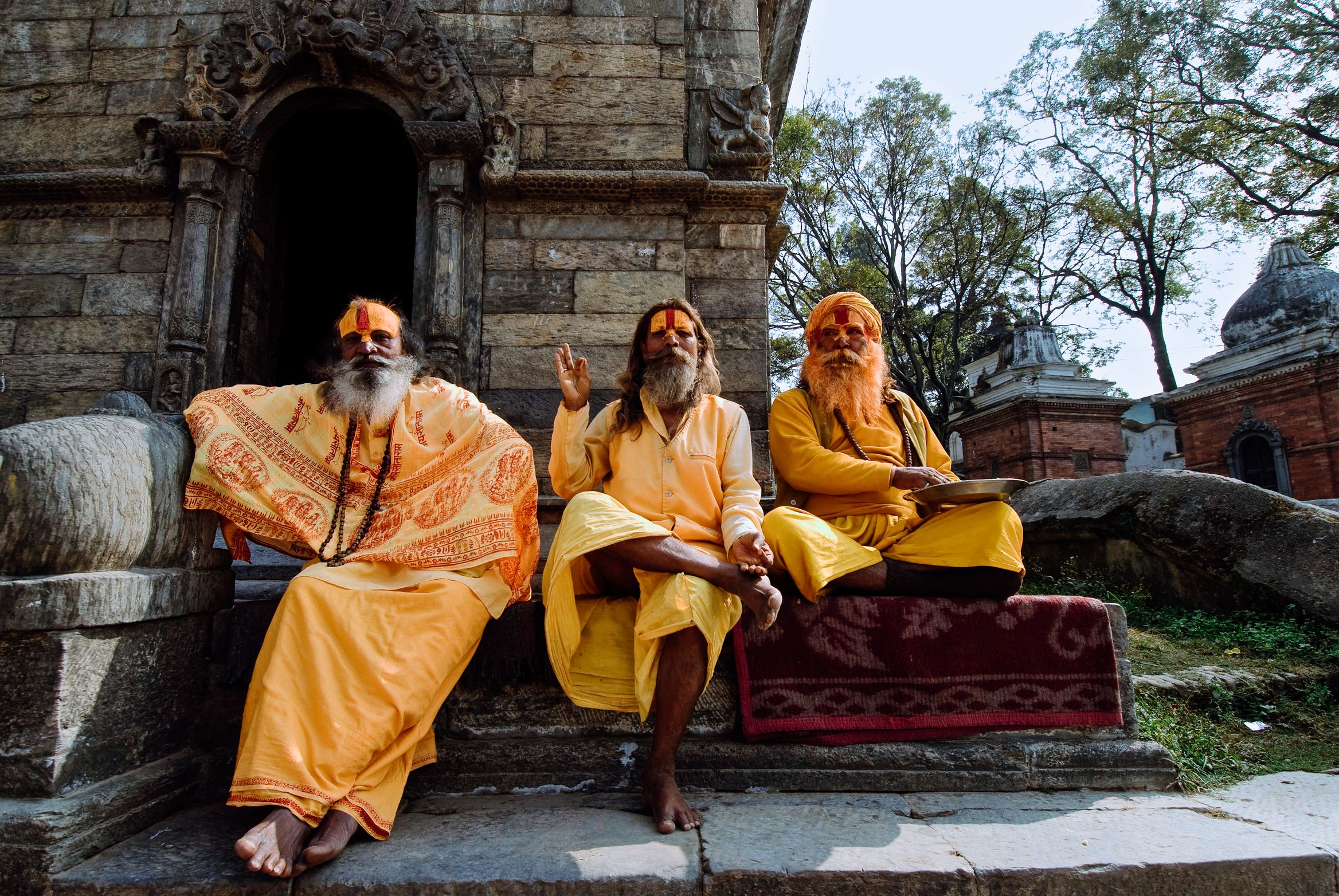 Three sadhus in saffron robes sitting on a shrine step, faces painted, one raising a hand mid-sentence