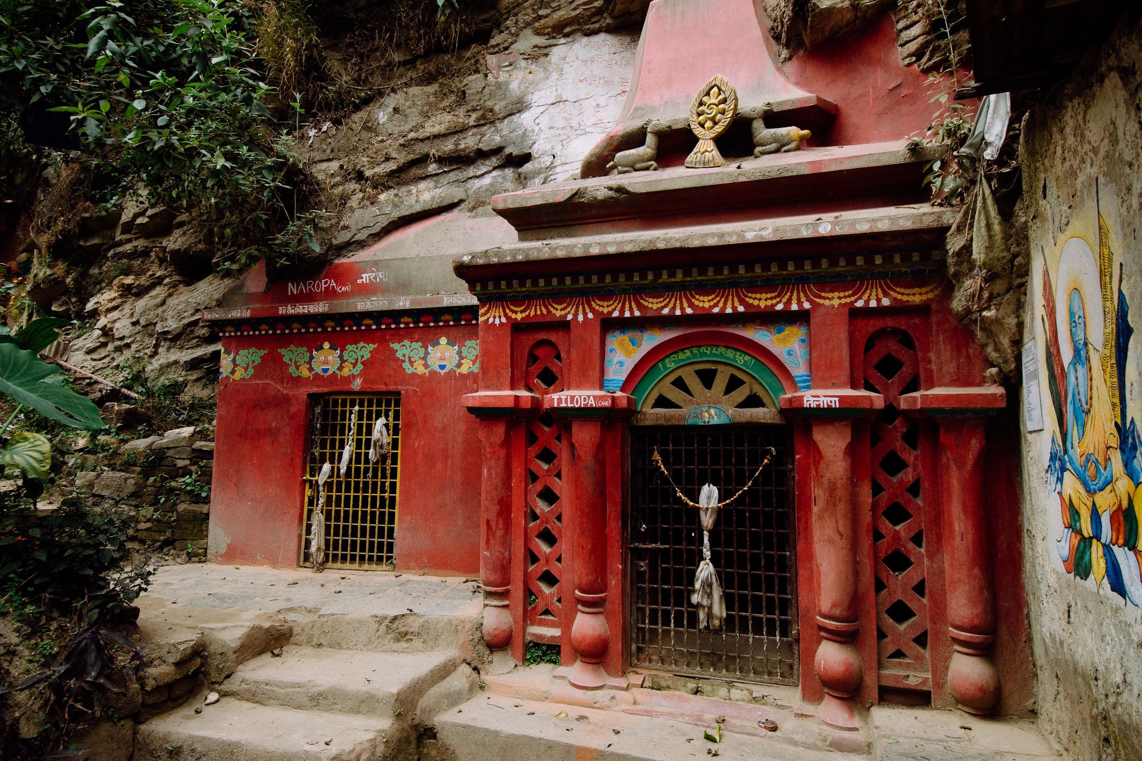 A bright red painted shrine built into the base of a cliff, a wheel of dharma above the door