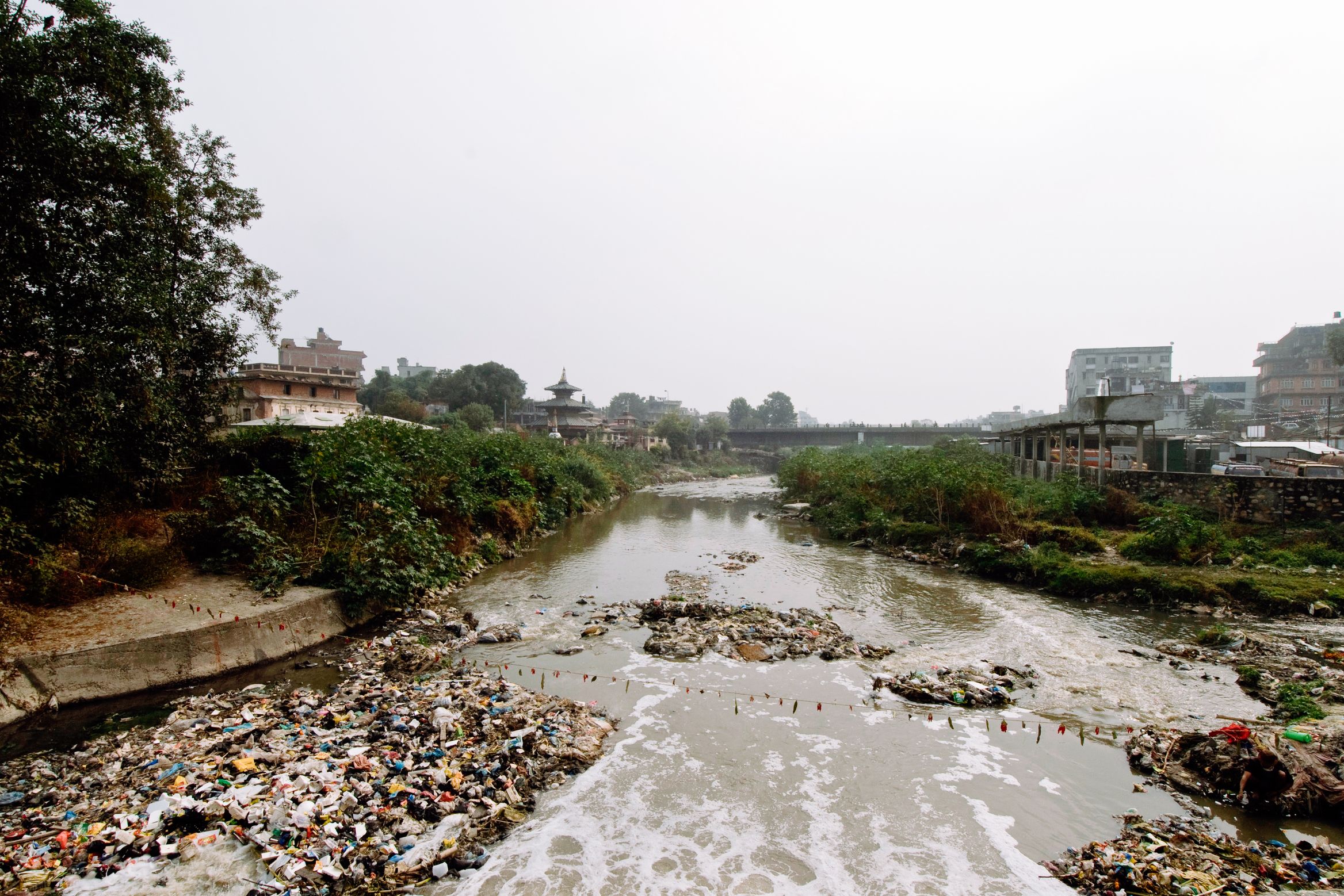 A river running white through banks of rubbish, town and temple roofs in the haze beyond