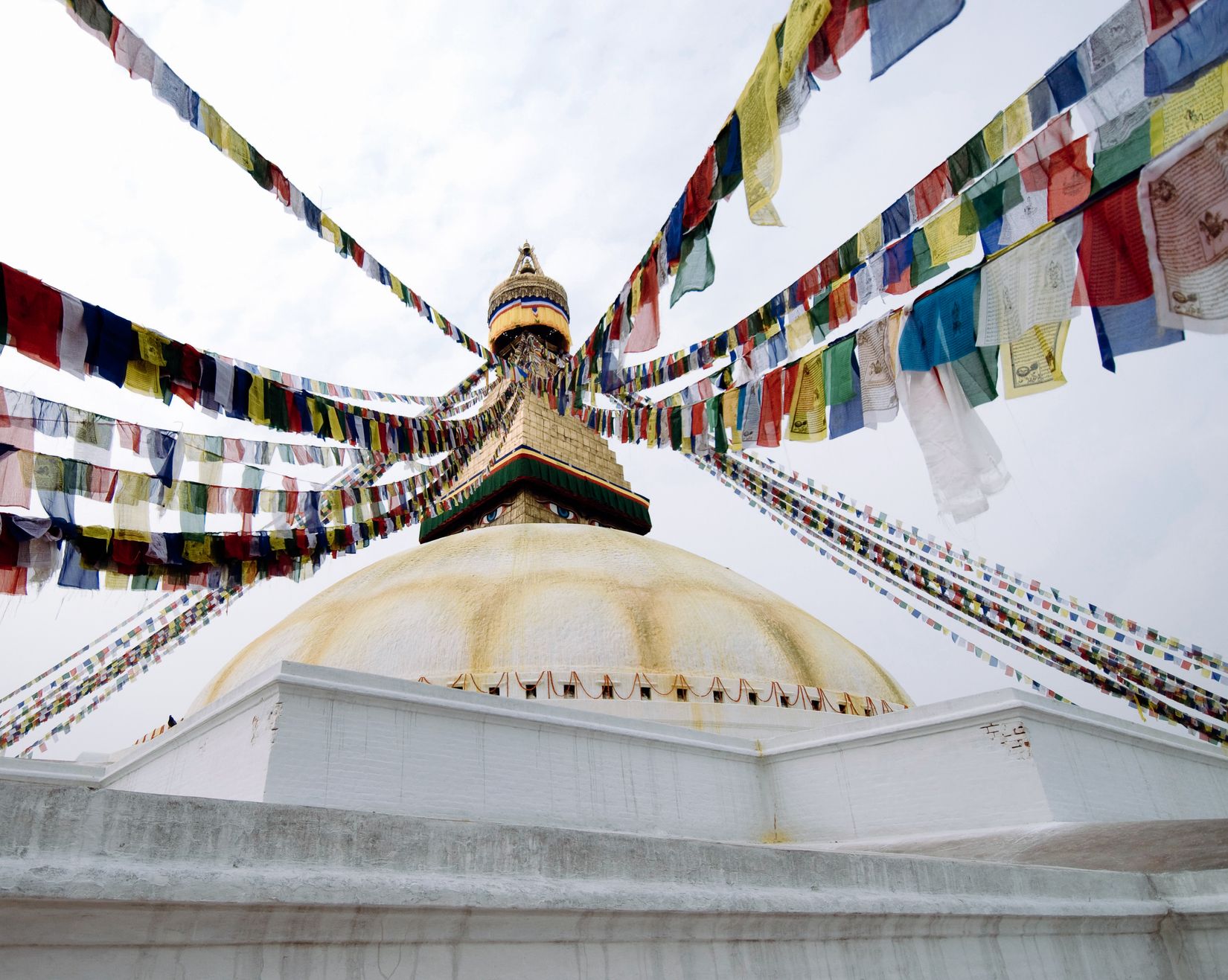 Looking up at the great white stupa, prayer flags radiating from the gilded spire across a blank sky