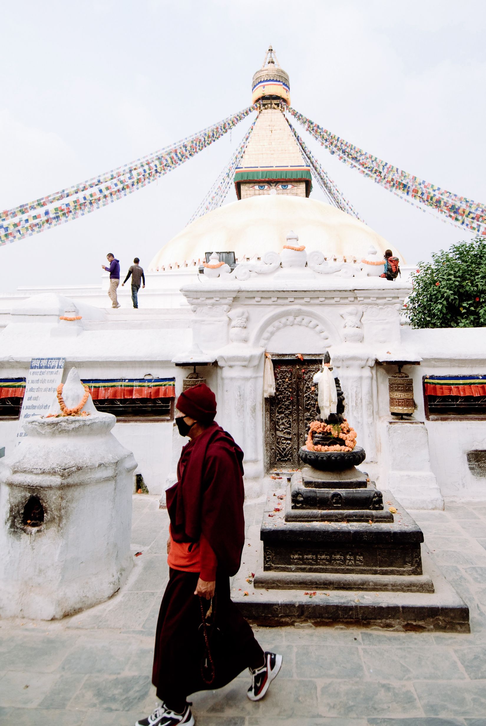 A monk in maroon walking the base of the stupa past a marigold-covered shrine