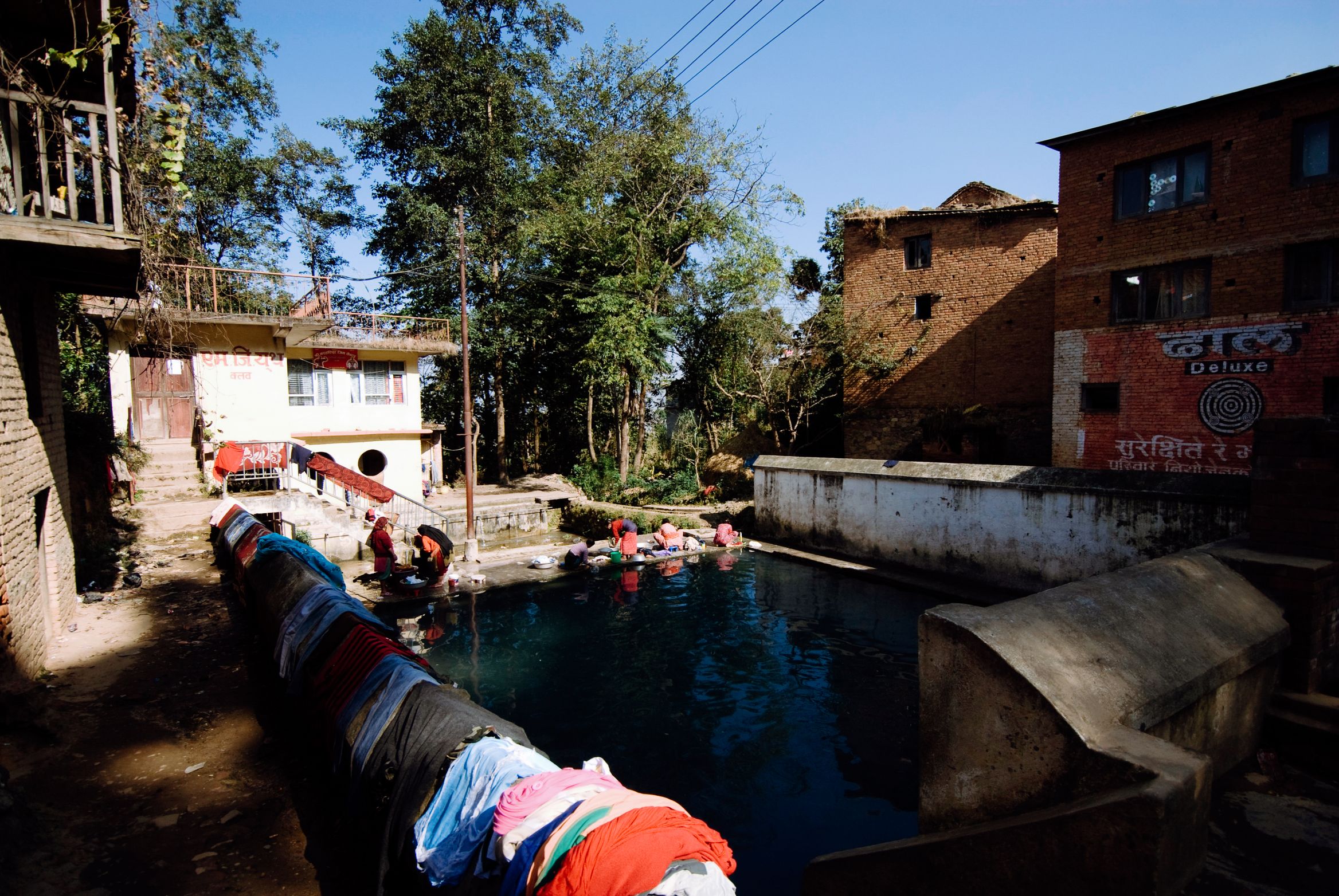 Washing spread along the wall of a village bathing pool, women in the water at the far end