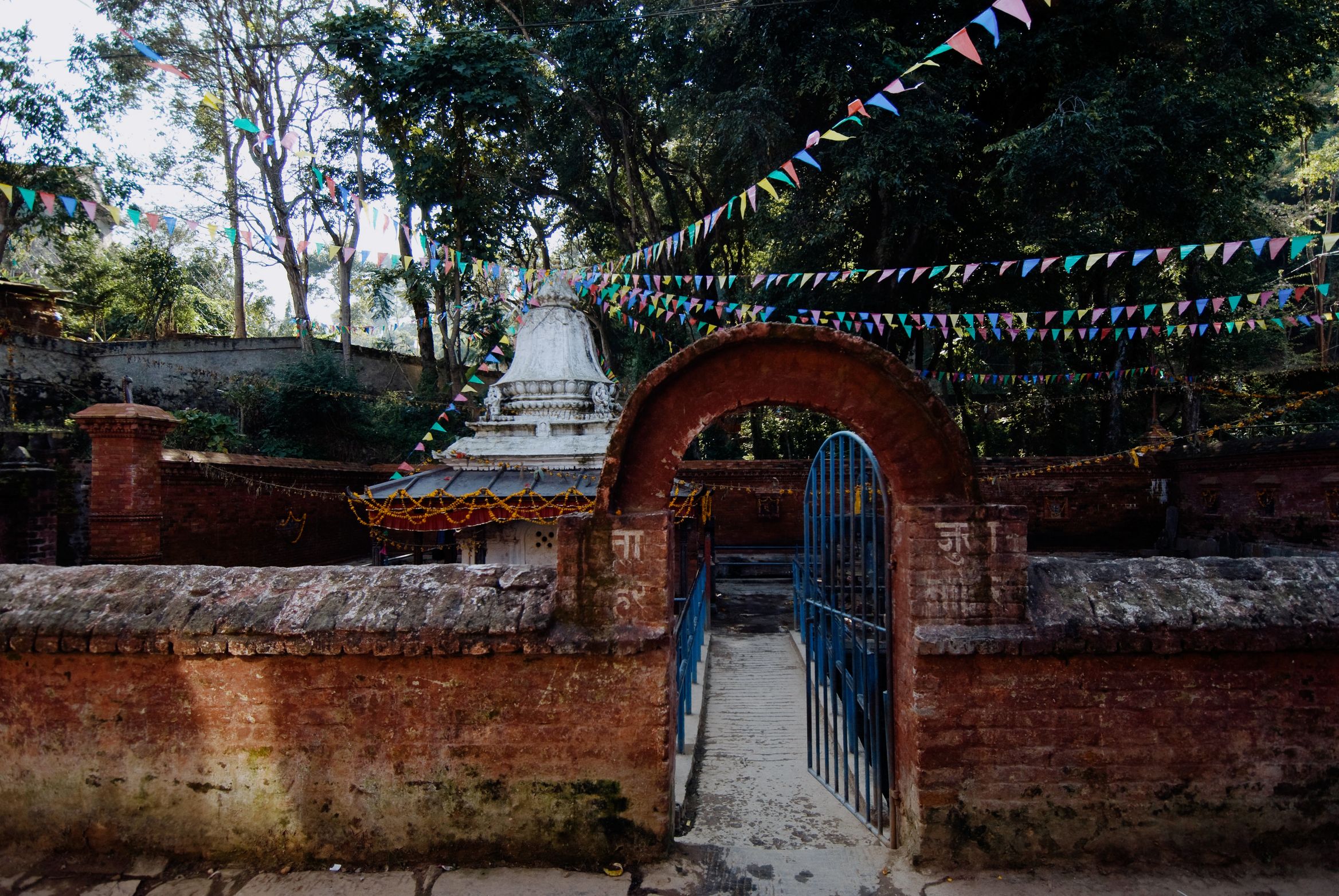 A brick gateway with a blue gate and a small white stupa behind, coloured bunting overhead