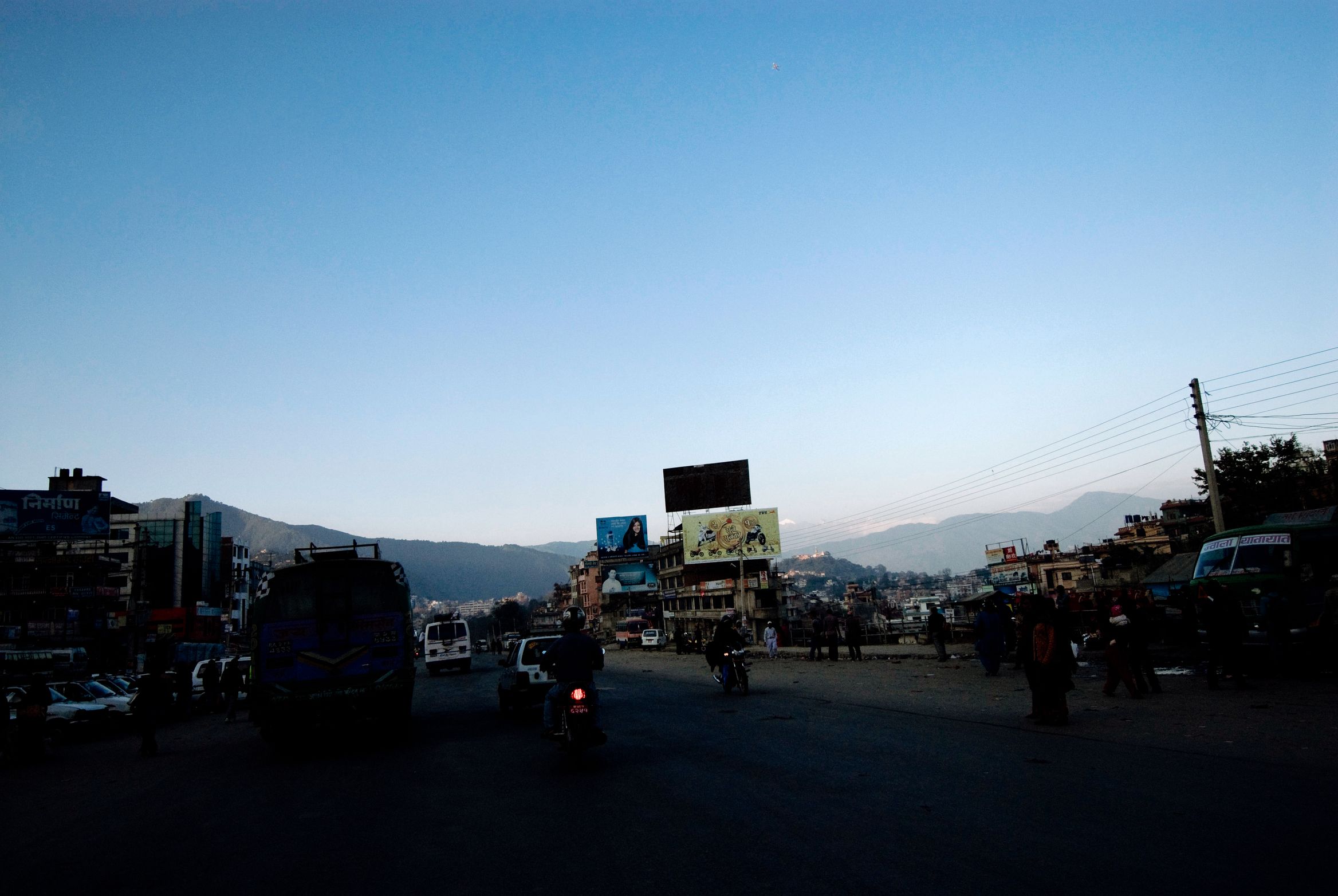 A wide dusty crossroads at dusk, buses and motorbikes silhouetted under a deep blue sky