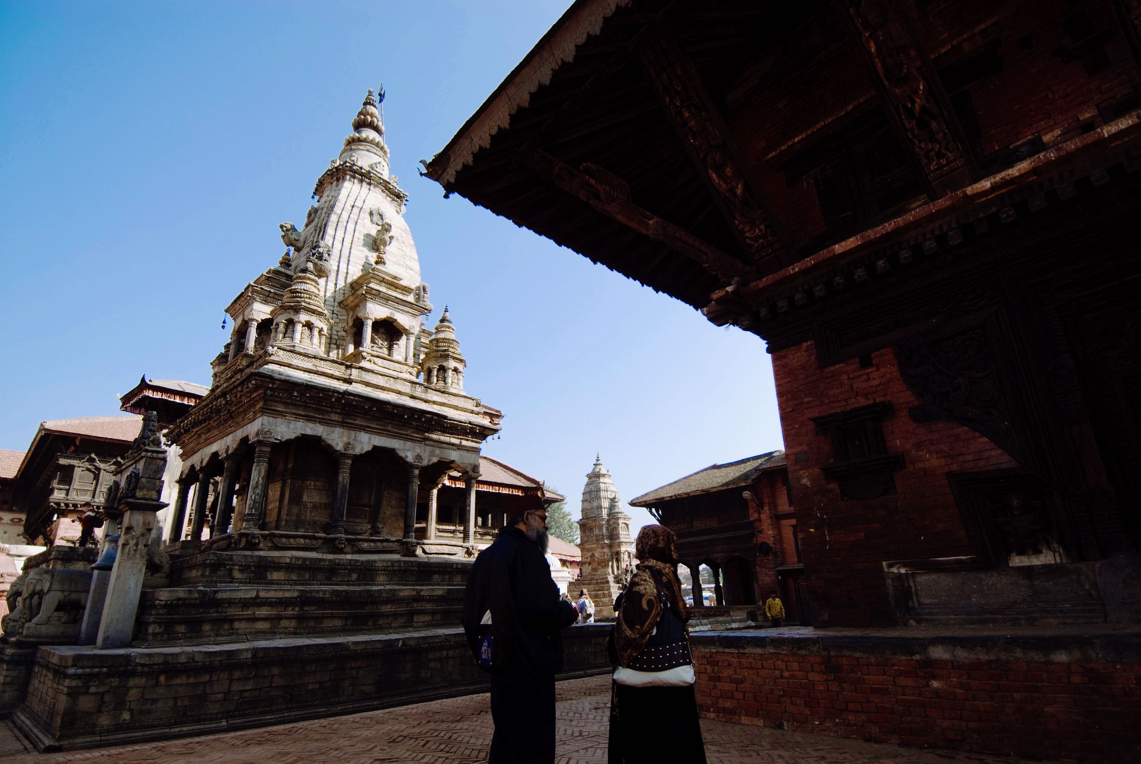 Two people standing in the shade of a temple eave, looking out at a white shikhara temple in the sun