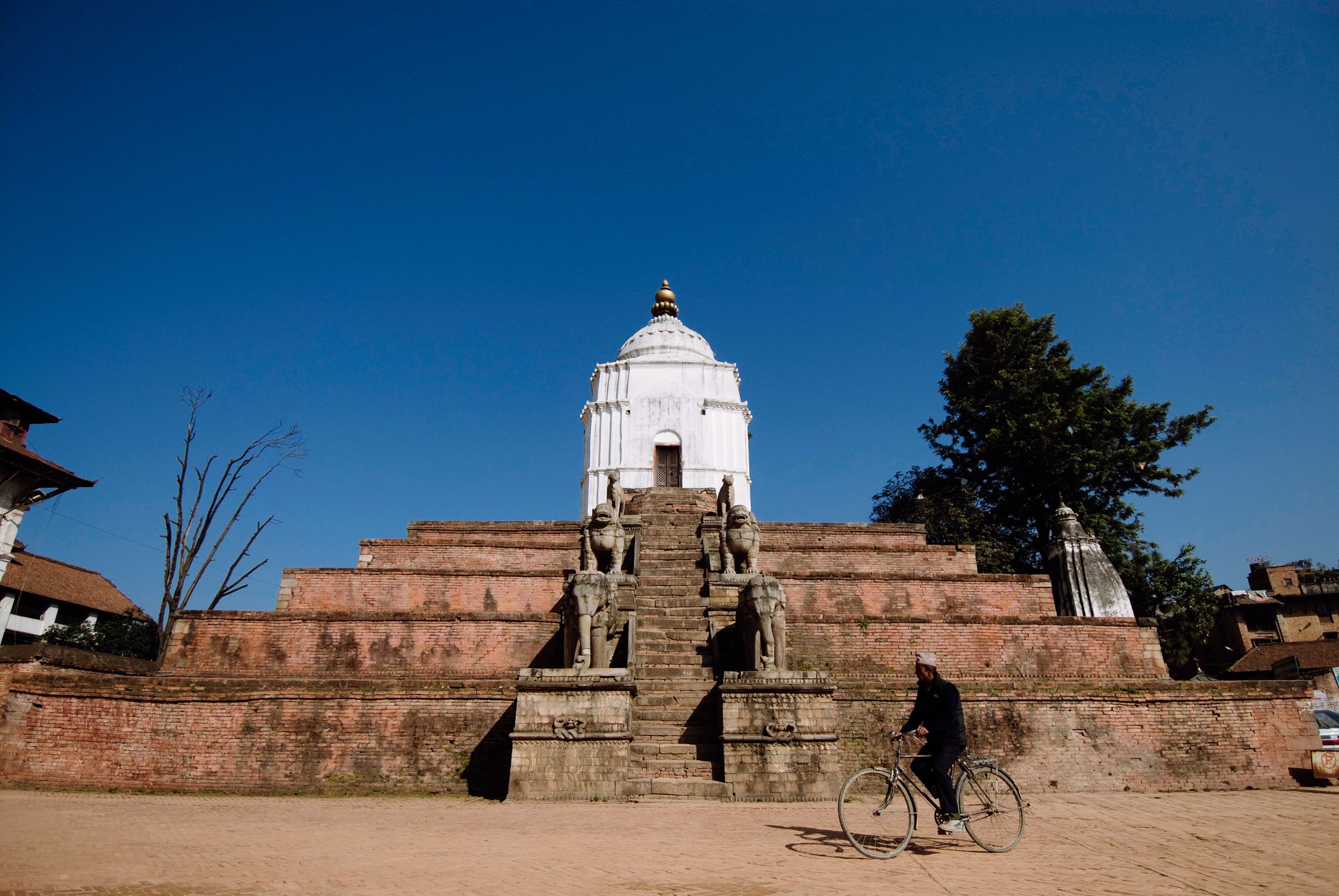 A man cycling past the stepped plinth of a white temple, sky hard blue above it