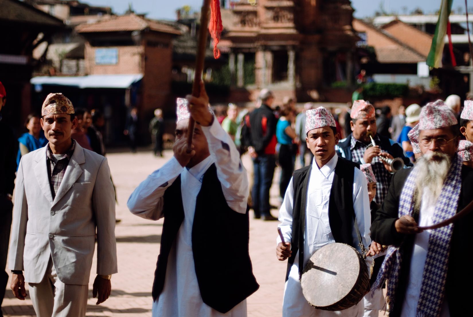 Men in topi hats processing through the square, one carrying a drum, another swinging a mallet