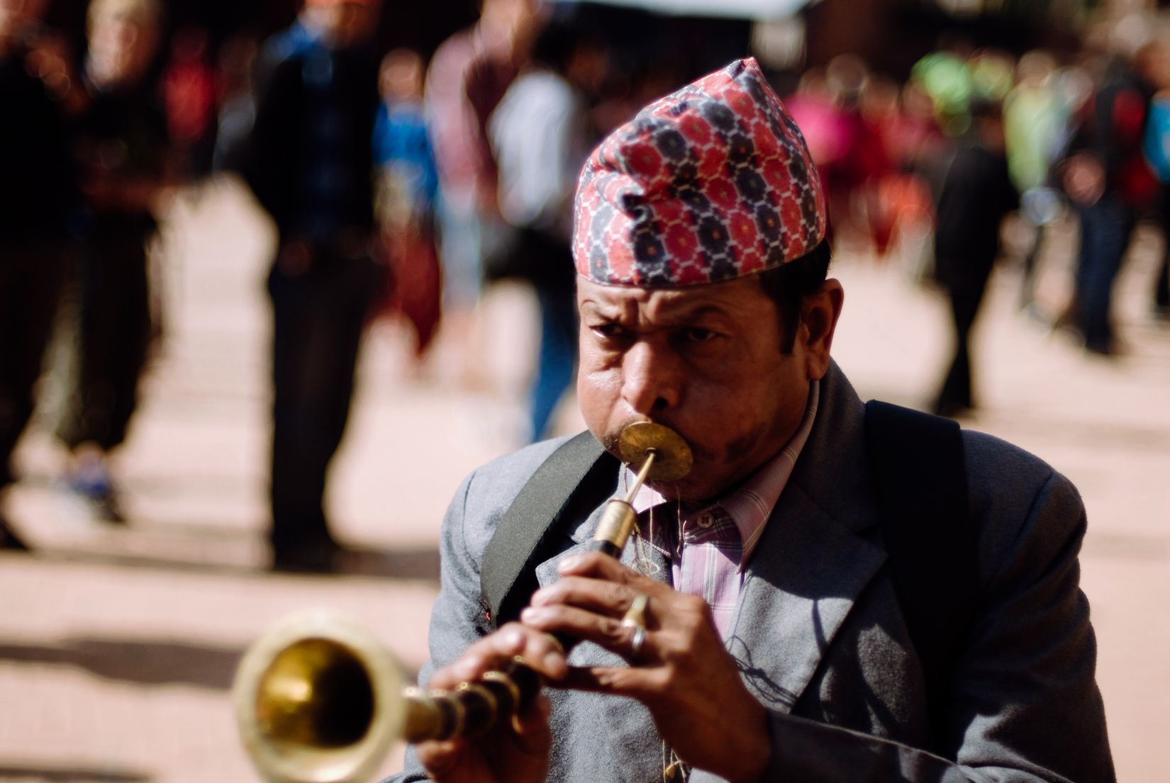 A man in a patterned topi playing a brass shehnai, cheeks full, crowd blurred behind him