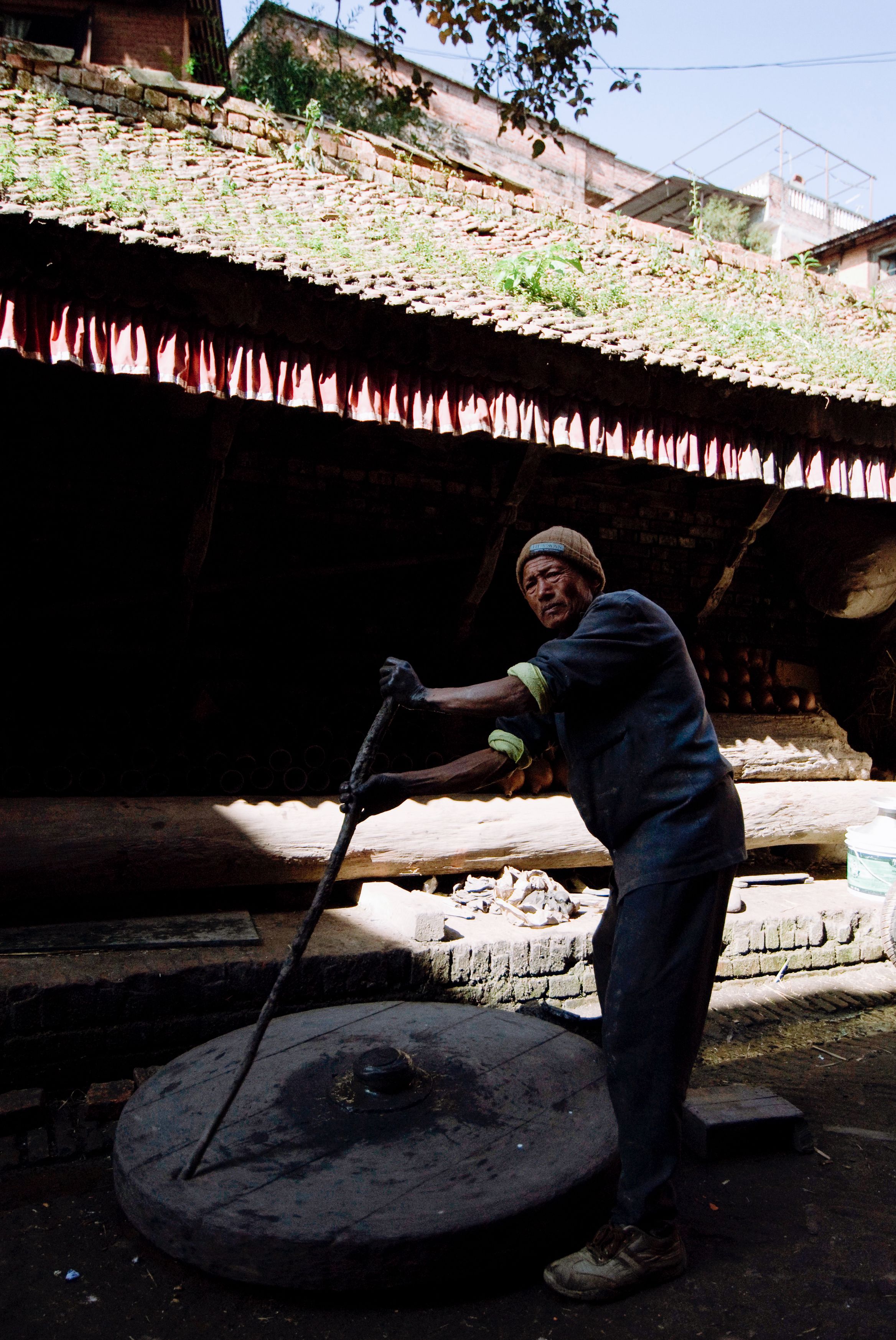 A man leaning on a stone grinding wheel in a courtyard, dyed red cloth drying on the roof above