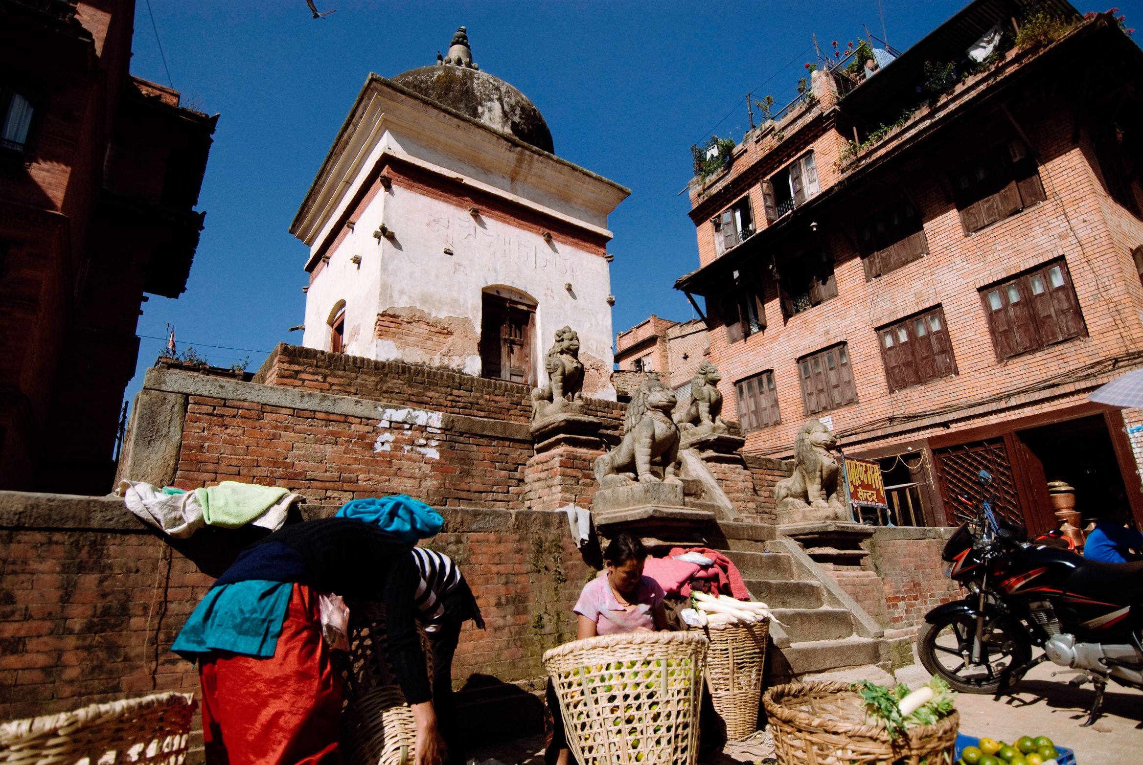Traders packing baskets of vegetables at the foot of a shrine guarded by stone lions