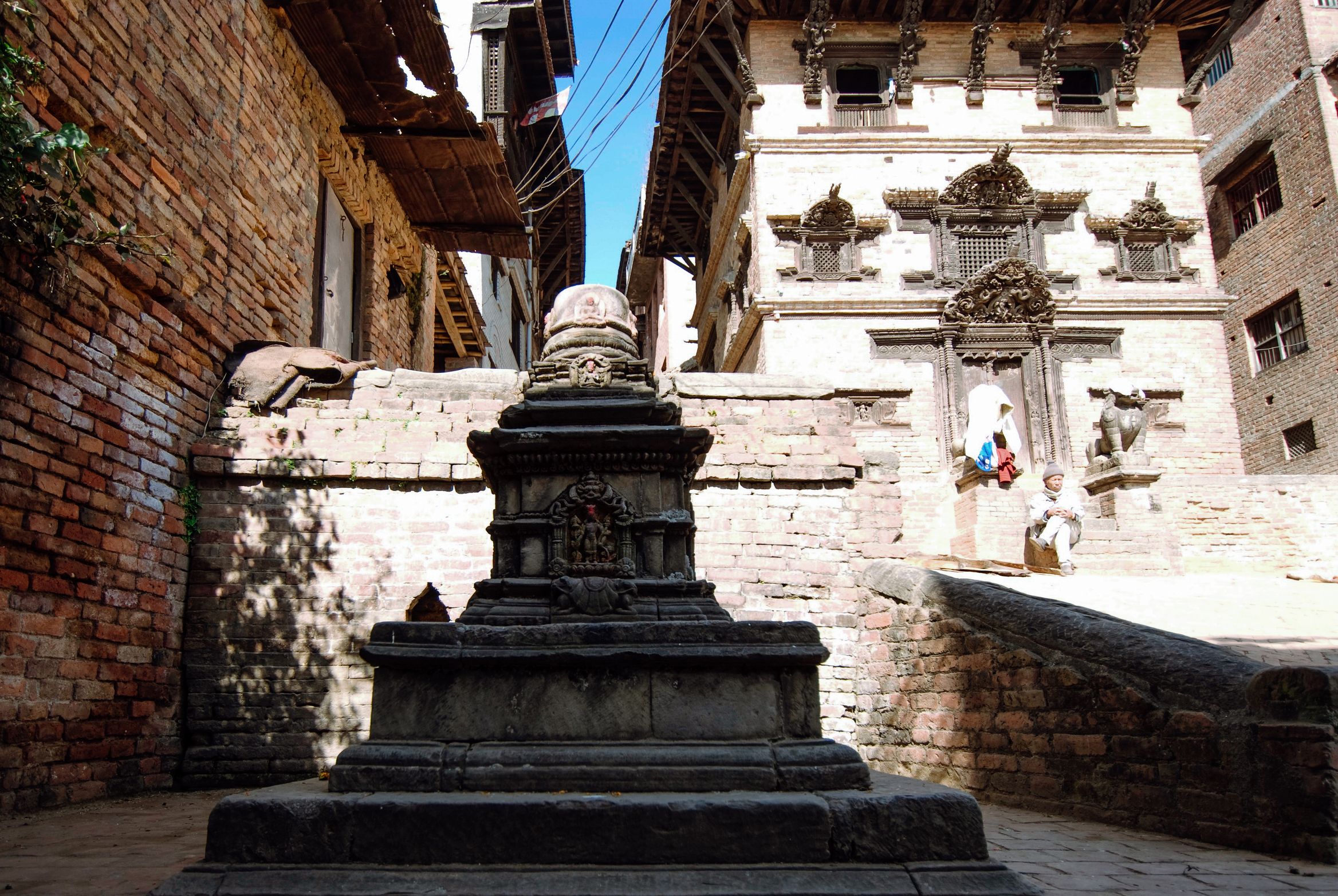 A dark stone stupa at the end of a narrow lane, a carved white house front catching the sun beyond