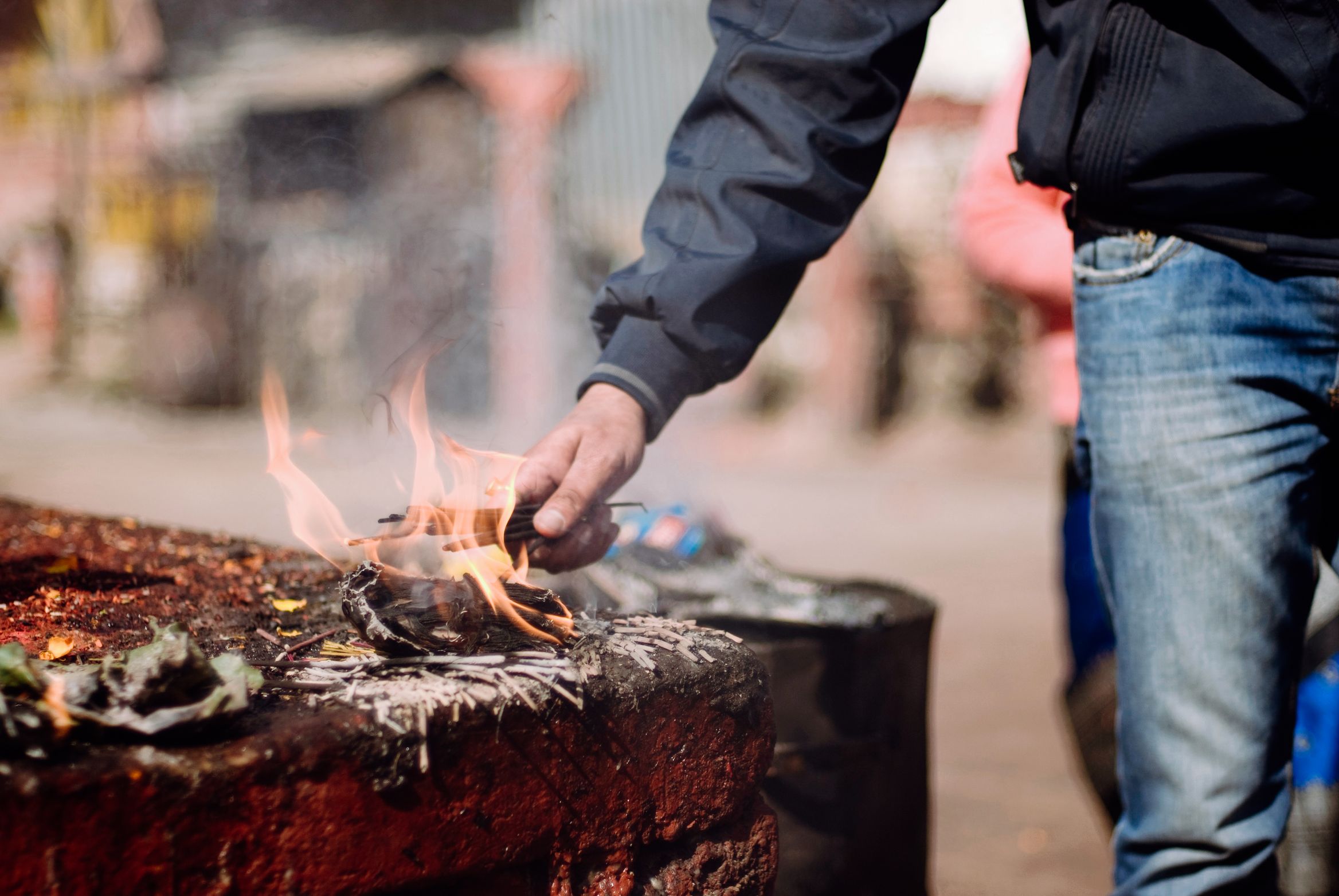 A hand setting light to an offering on a red-washed stone platform, flames going up pale in daylight
