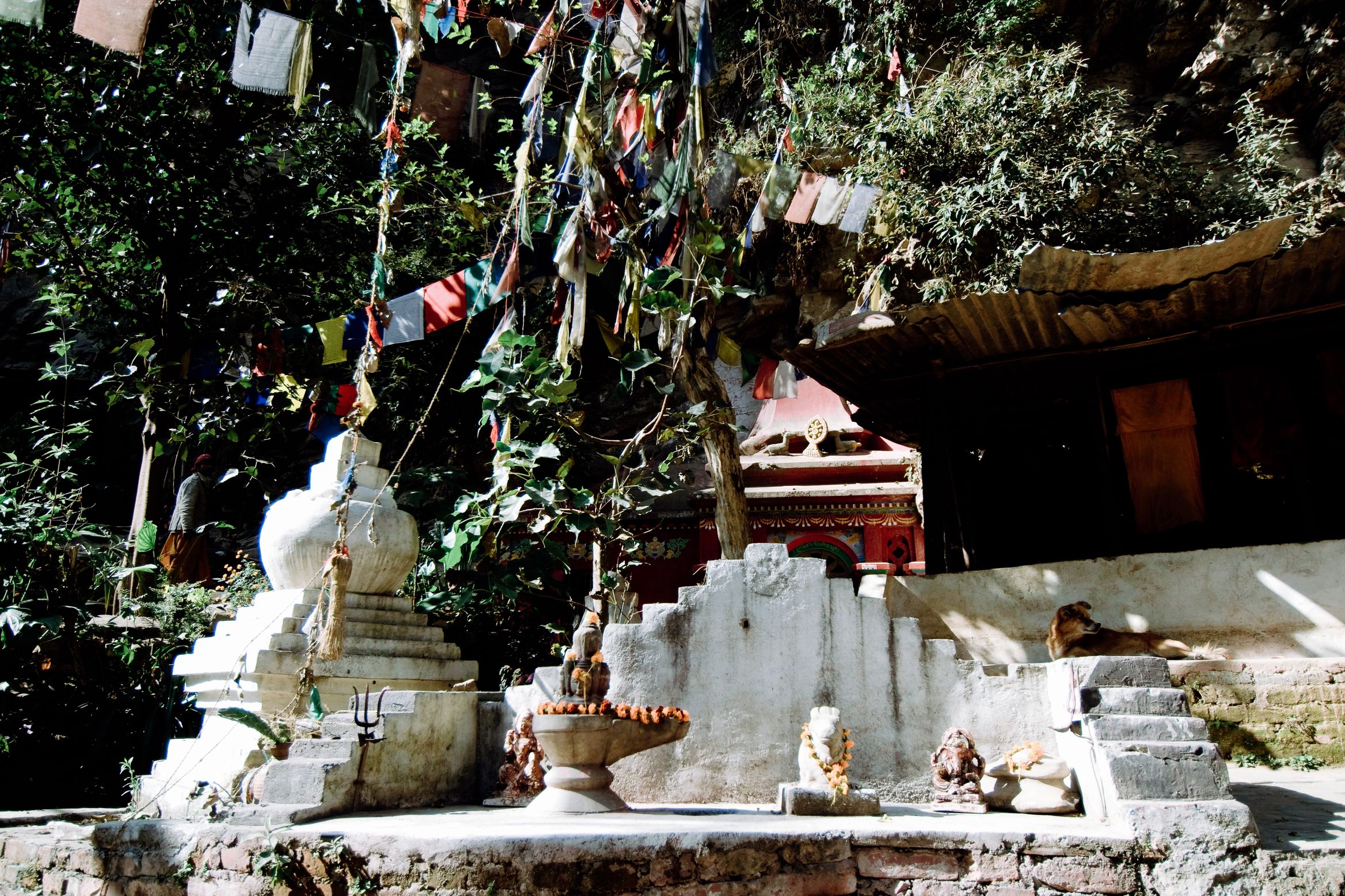 A shrine under a cliff hung with faded prayer flags, a white stupa and offering bowls in front