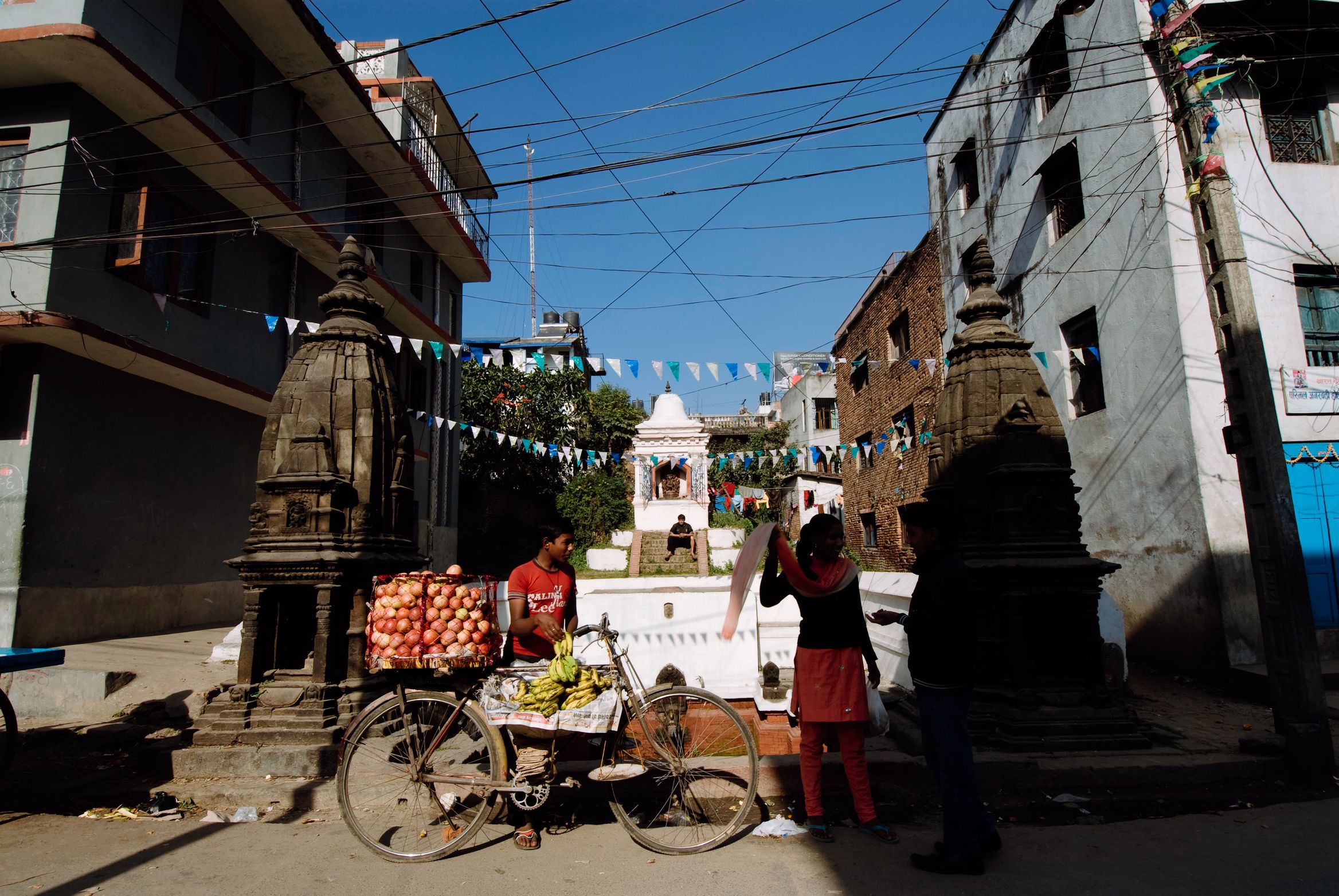 A fruit stall on a bicycle parked by a white shrine at a bunting-strung street corner