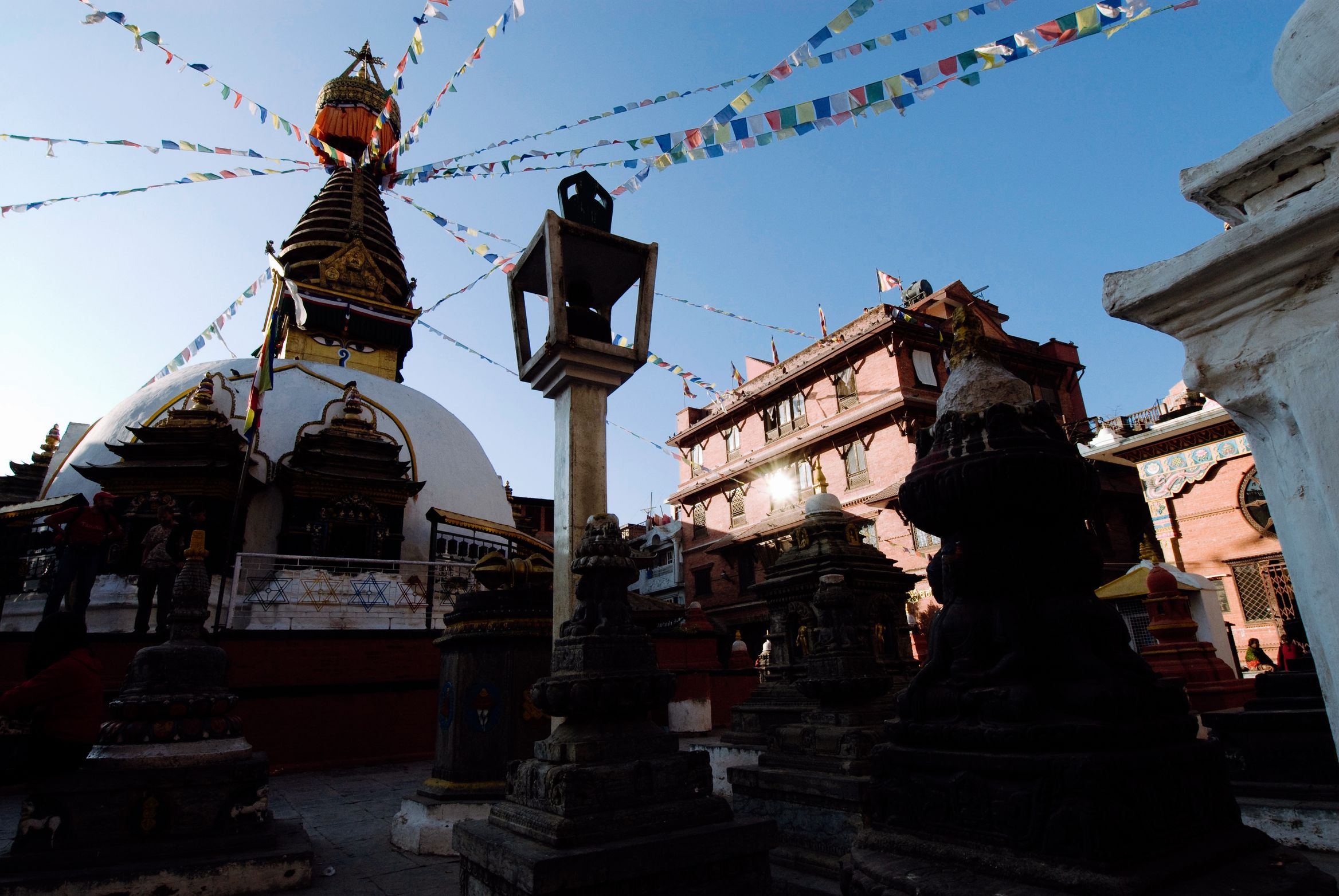 A white stupa and stone shrines at dusk, prayer flags fanning out overhead against a darkening sky