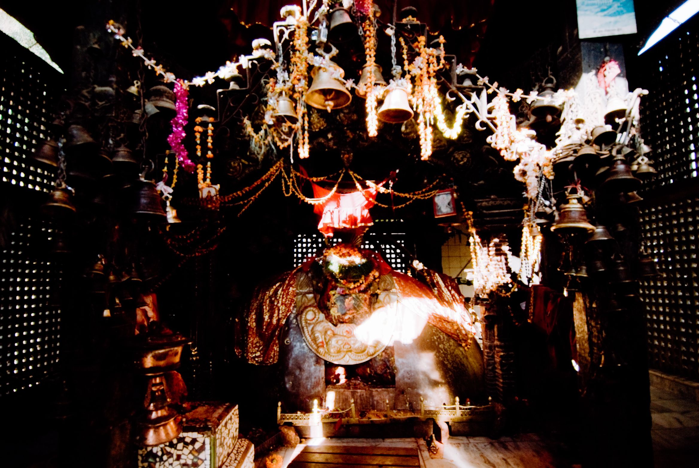 The inside of a temple hung thick with brass bells and offerings, a lamp burning at the shrine