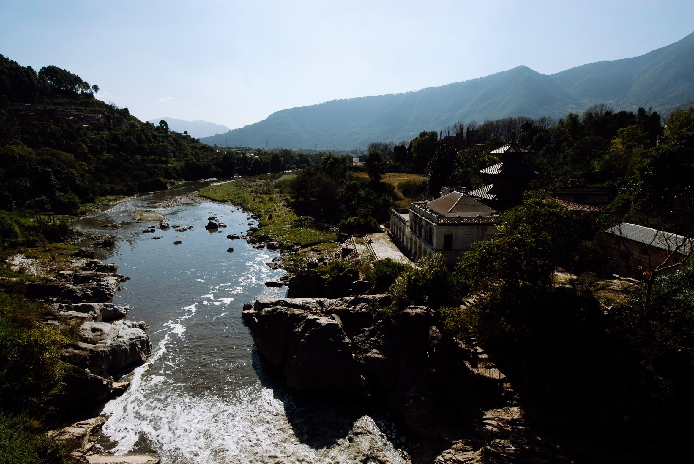 A river breaking white over rocks below a road, wooded hills rising on both sides