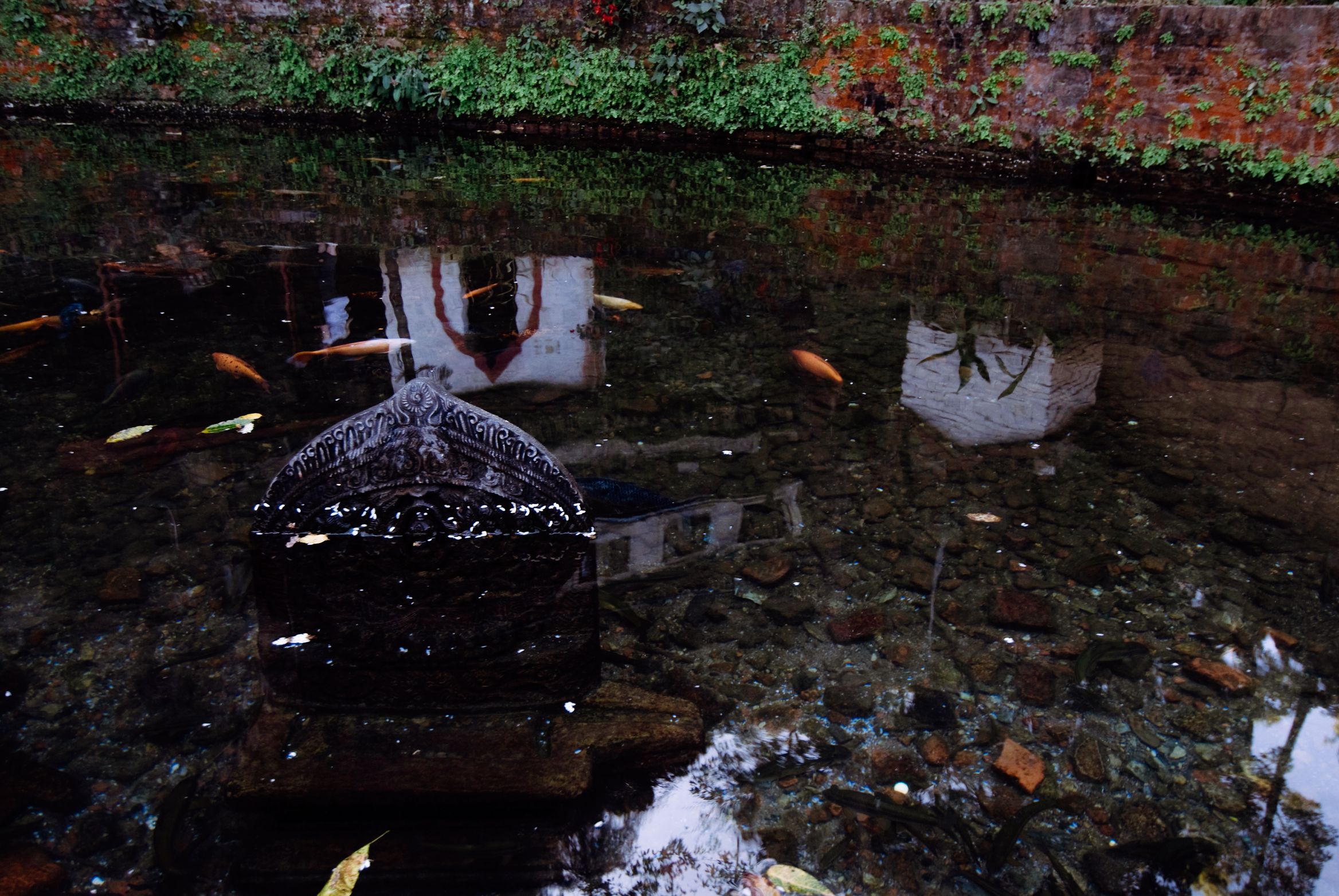 A small stone shrine standing submerged in a shallow green tank, brick wall reflected on the surface