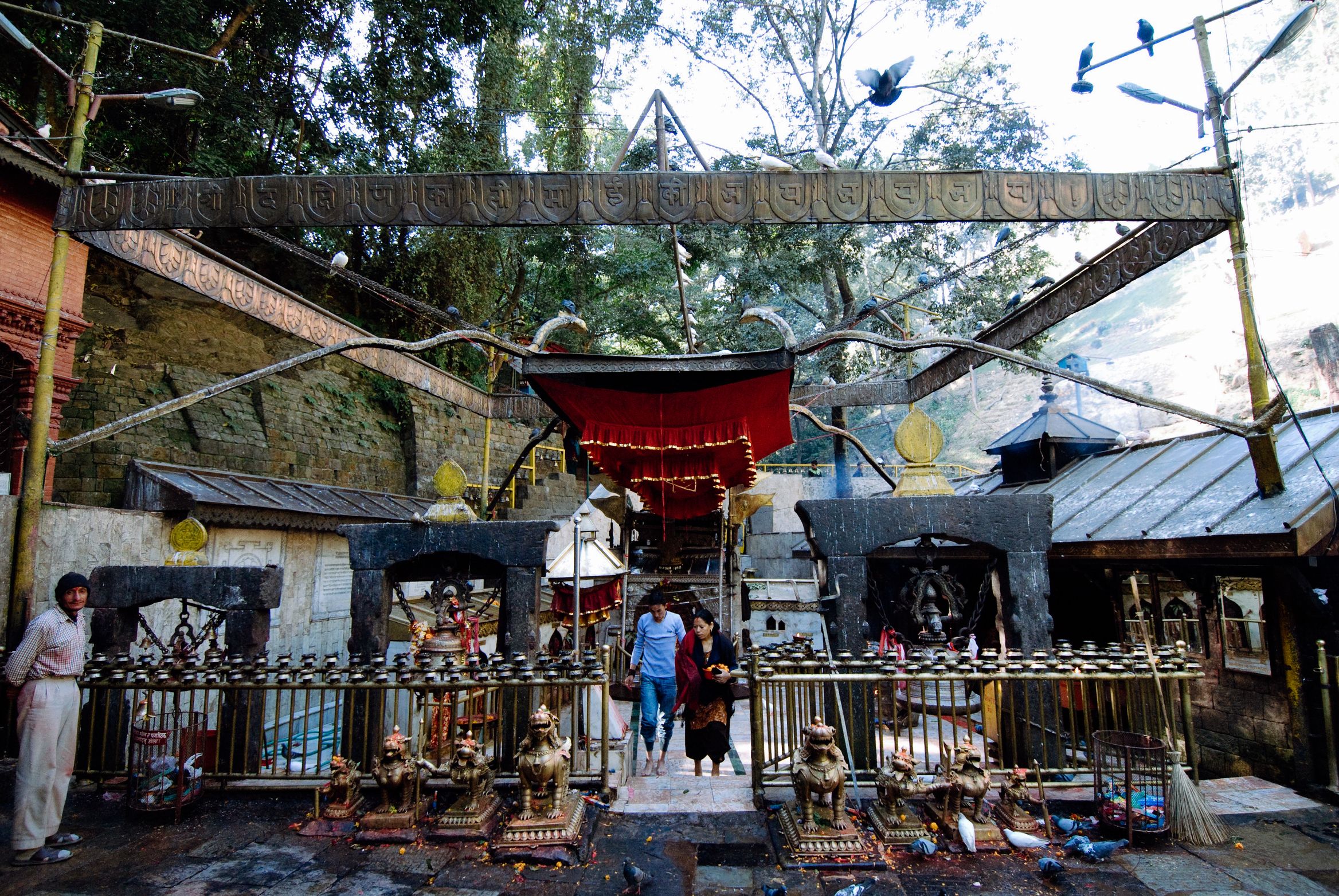 A red-roofed shrine behind railings crowded with trident offerings, two people standing at the gate