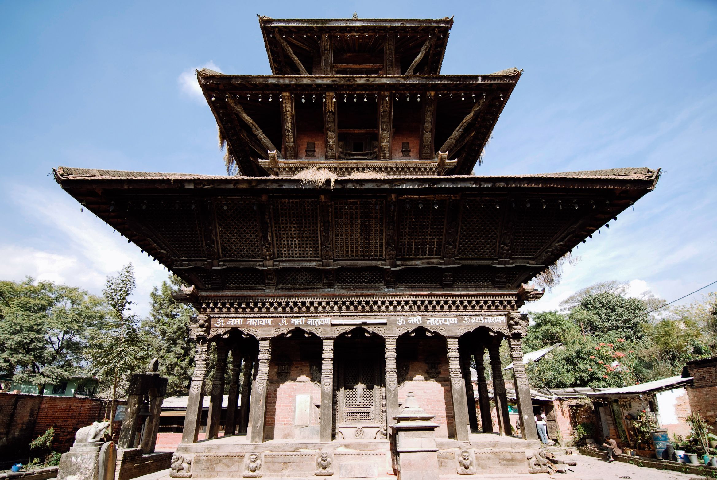 A three-tiered wooden temple seen straight on, carved struts under each roof, empty in the sun