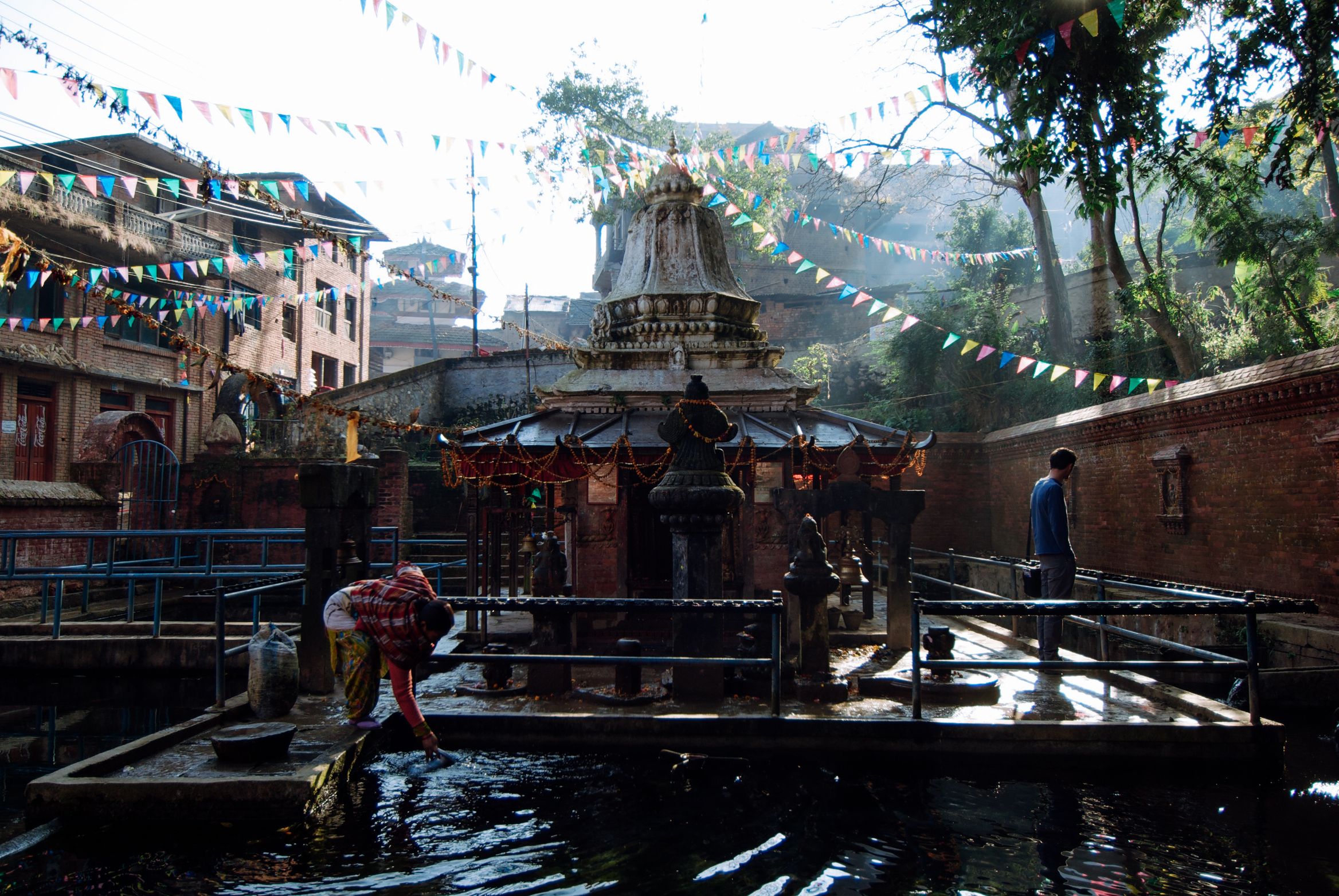 A shrine standing in a village water tank under bunting, a woman in red carrying a load down to the edge