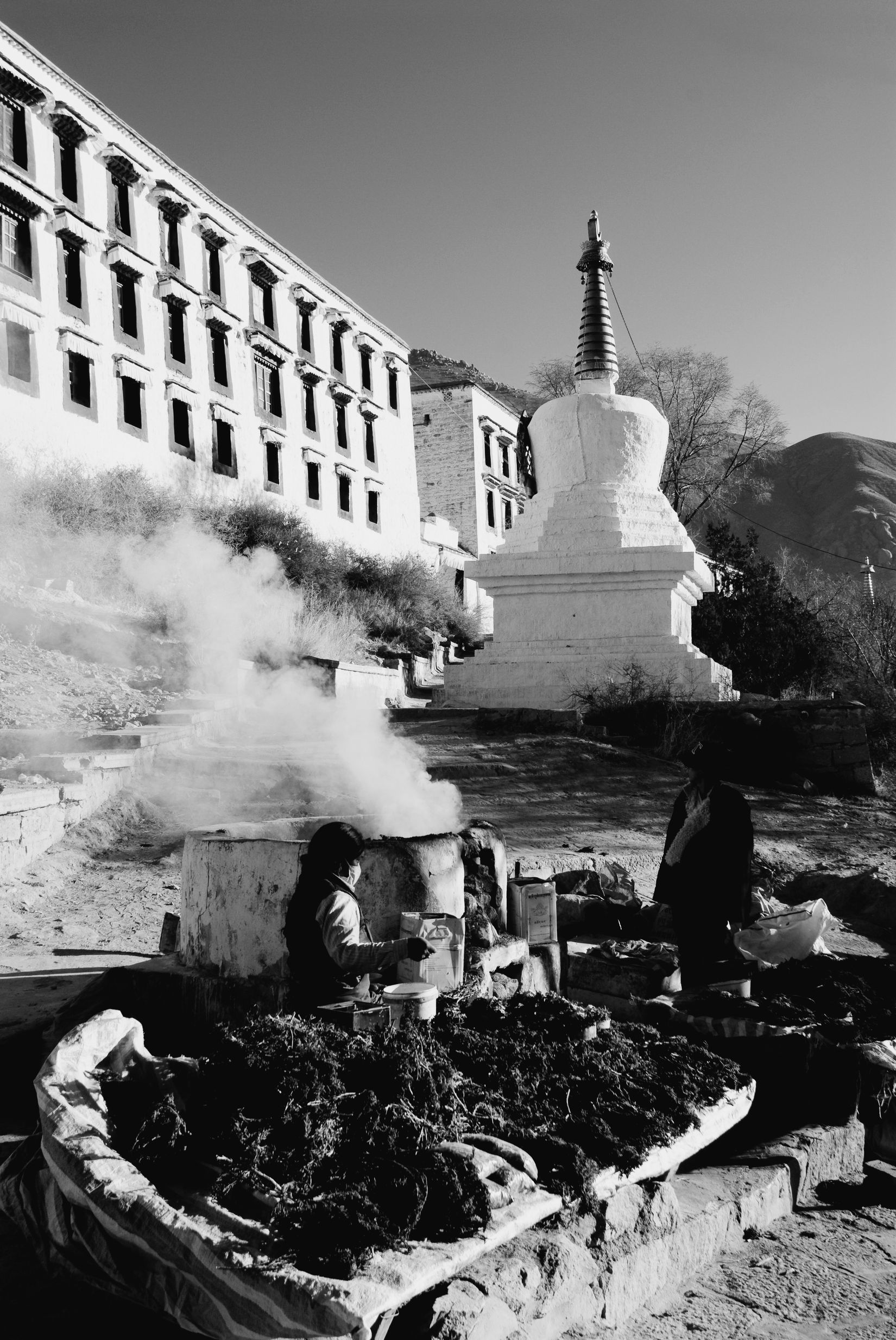 Black and white: juniper smoke rising by a white stupa below the monastery buildings