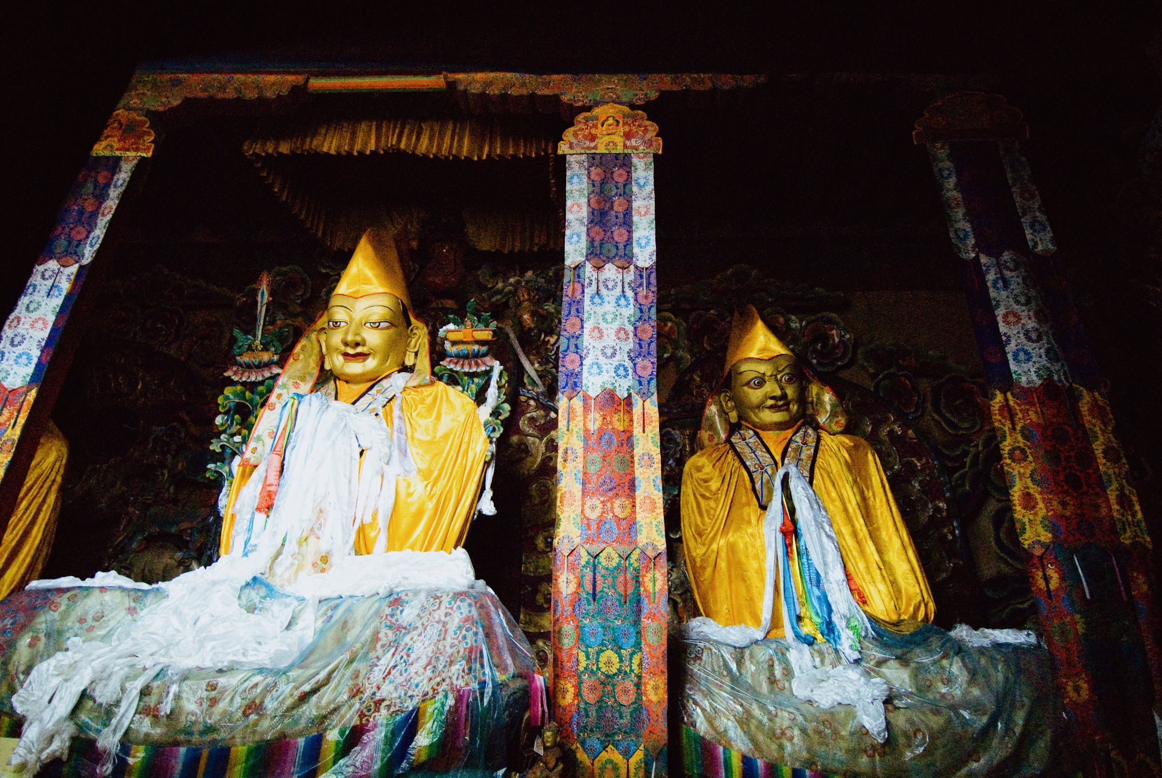Two gilded figures in yellow hats inside a dim chapel, draped with white offering scarves