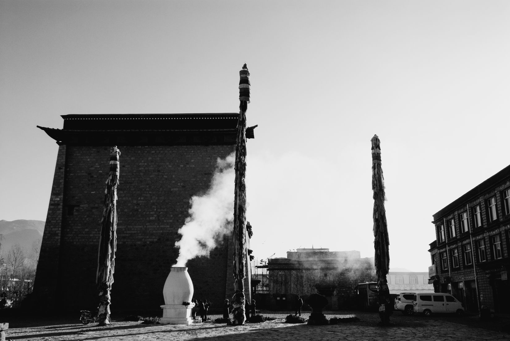 Black and white: incense smoke from a white burner between tall prayer poles by a monastery wall