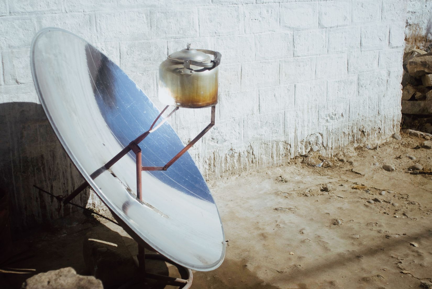 A parabolic solar cooker propped against a wall with a kettle at its focus