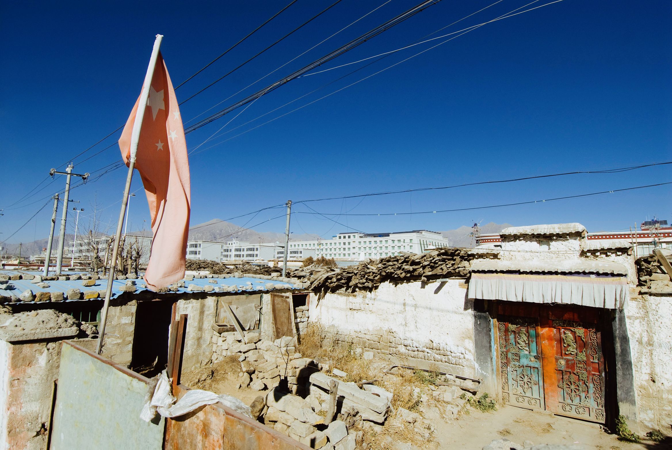 A flag on a pole over a whitewashed village house with a painted wooden door, power lines overhead